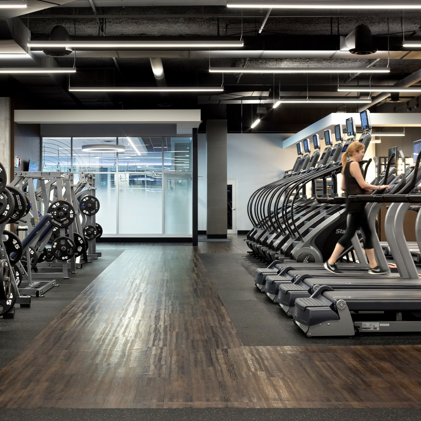 Interior of a modern gym with weight lifting equipment on the left and a row of treadmills on the right, a person walking on a treadmill, glass window in the background.