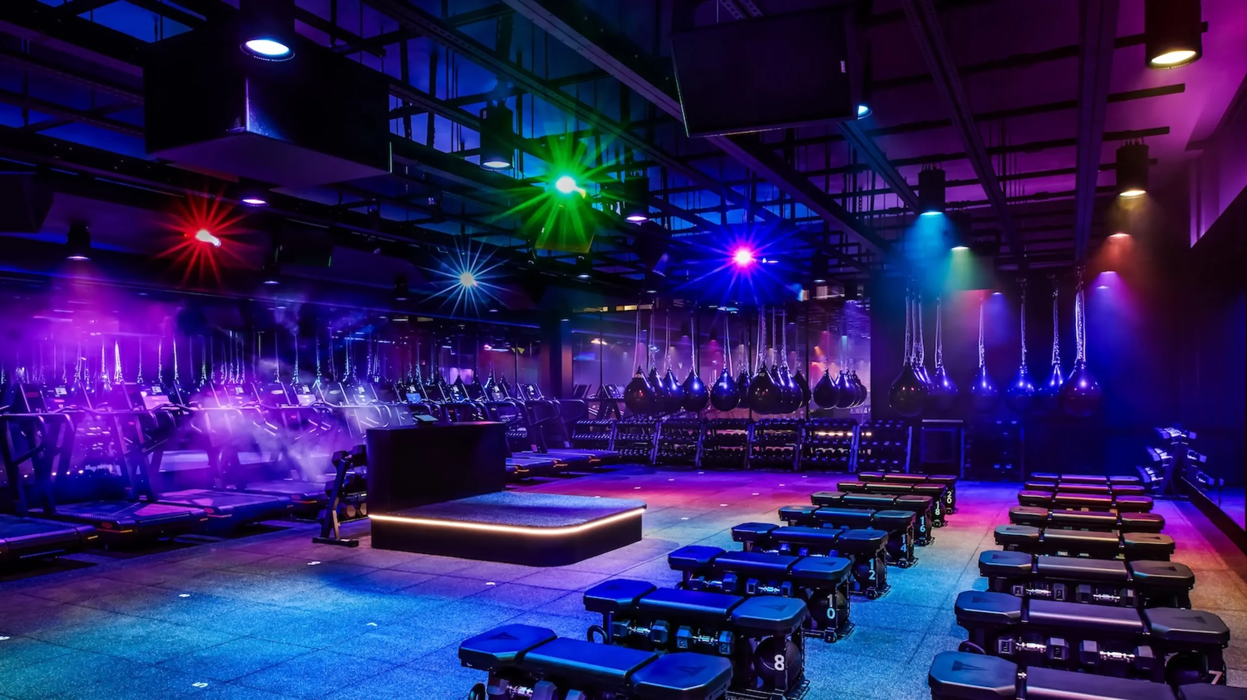 A dimly lit gym with colorful lights, exercise benches, and suspended punching bags.