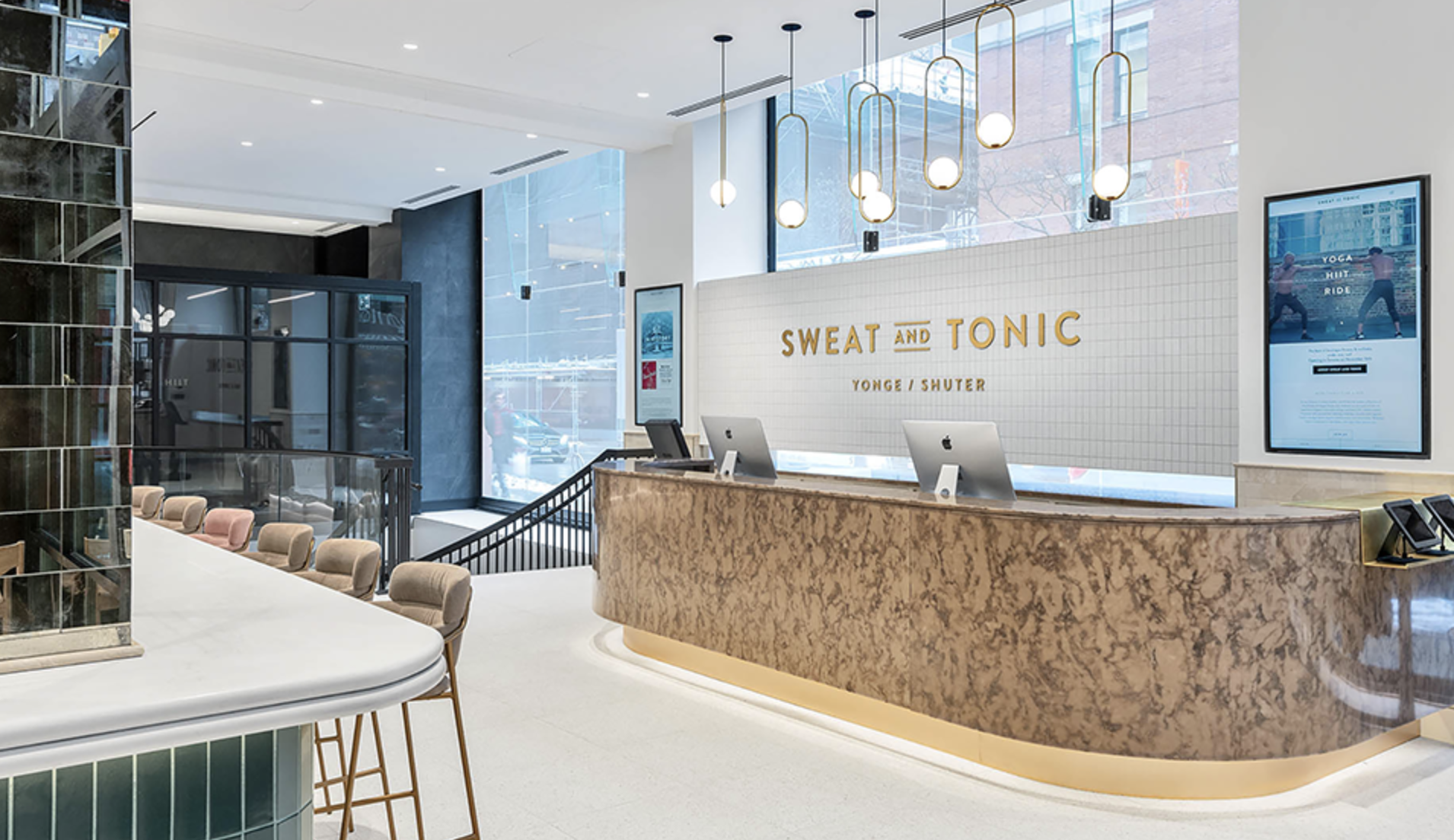 Hotel reception area with a marble front desk, digital check-in displays, and pendant lighting fixtures, with a seating area and large windows
