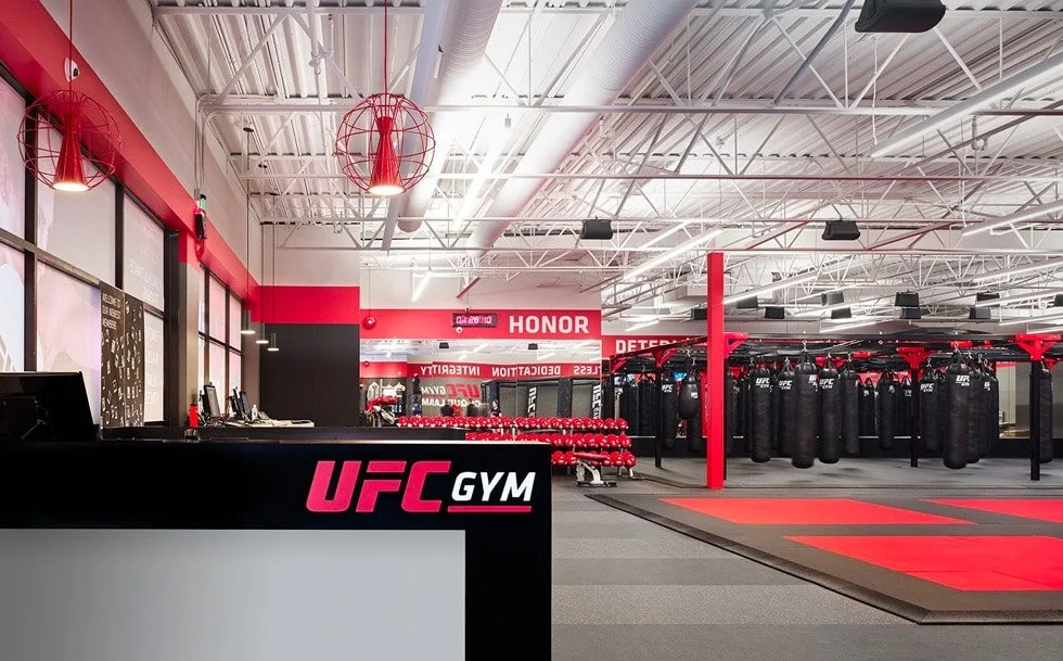 Interior view of UFC GYM with a front desk, red and black color theme, punching bags, a mat area for martial arts or workouts, and fitness equipment in the background.
