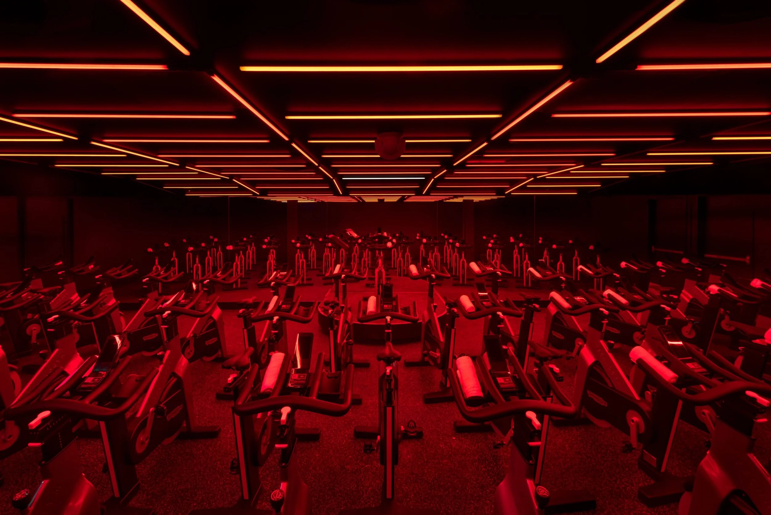 Indoor cycling studio with multiple stationary bikes under red and orange linear ceiling lights.