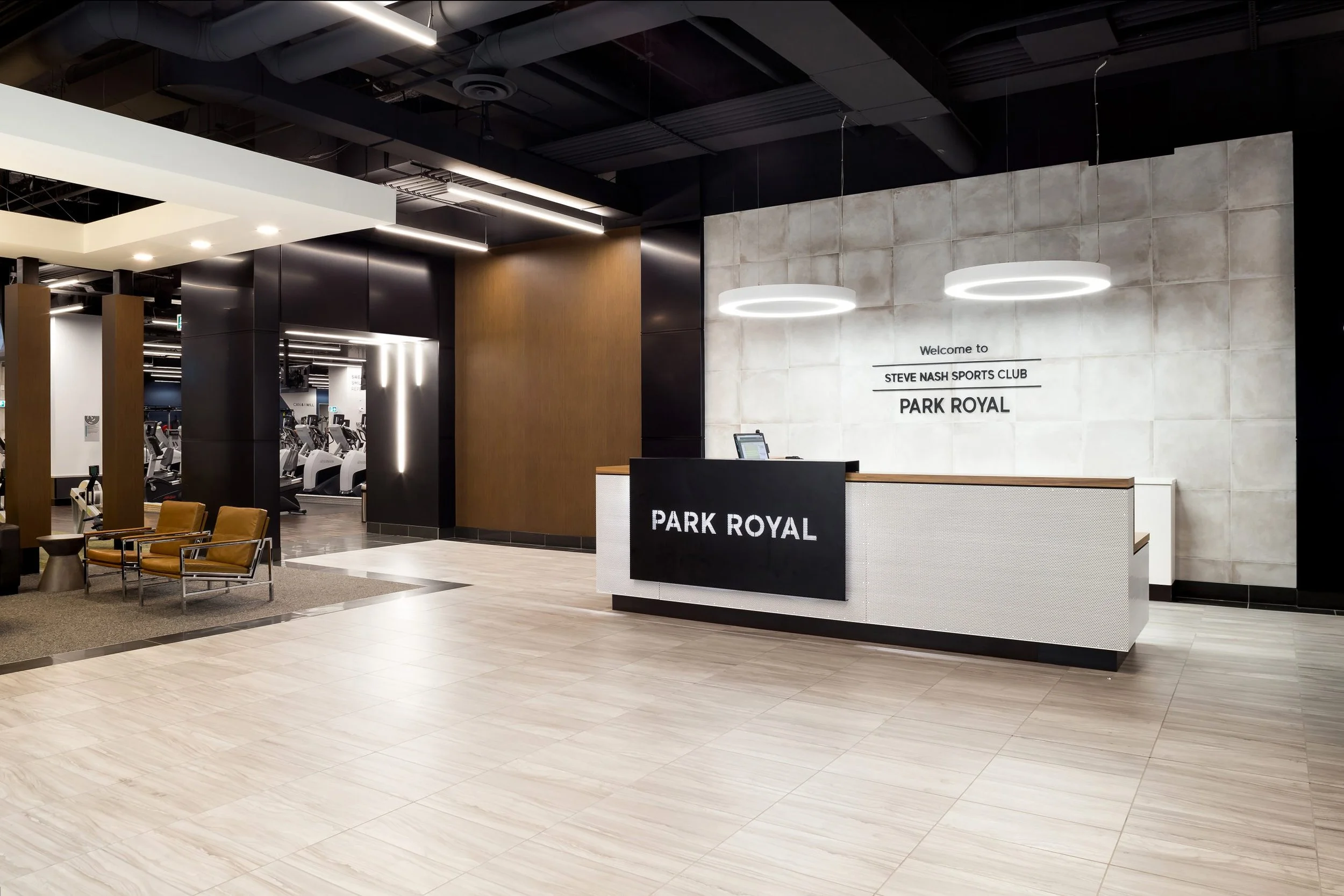 The reception area of Steve Nash Sports Club Park Royal featuring a modern design with a black reception desk, white tiled wall, and seating area with chairs. In the background, there are cardio machines in the gym area.