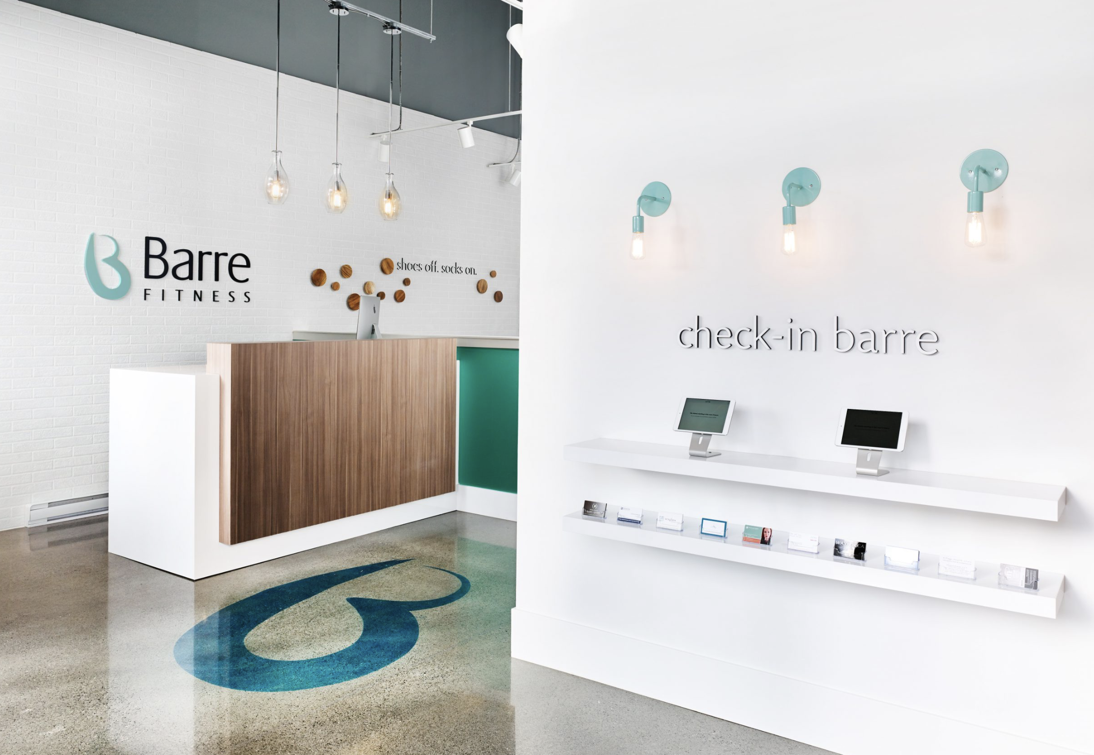 Interior of a fitness studio reception with a check-in area, modern lighting, and signage for Barre Fitness and check-in Barre, featuring a white wall, digital devices on a white shelf, and a logo on the wall.