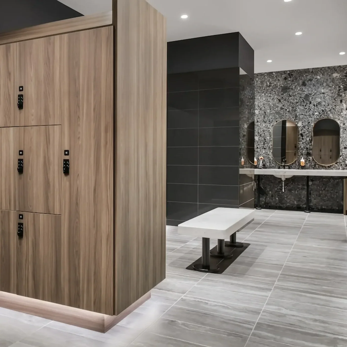 Modern bathroom with wood lockers, a white bench, dark tiled wall, granite patterned backsplash, oval mirrors, sinks, and fluorescent ceiling lights.