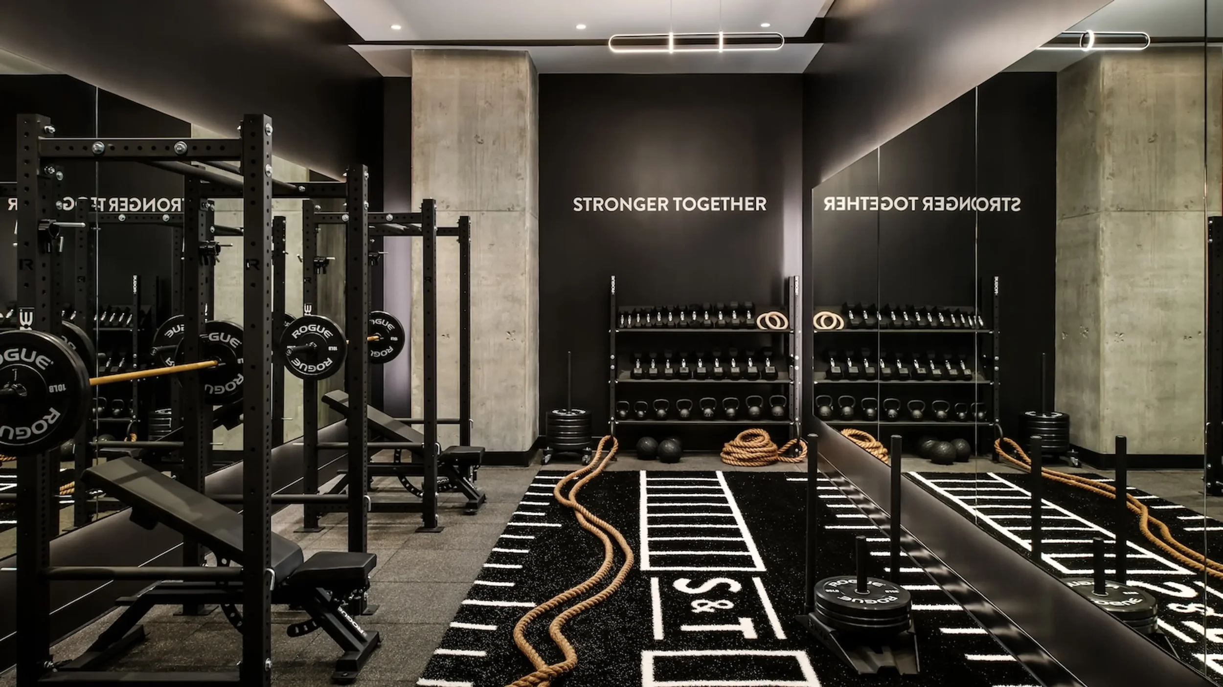 Modern gym with weightlifting equipment, including black shelves with dumbbells, barbells, and rubber medicine balls. Ropes are on the floor, and a black mirror wall reflects the room. The wall has the text "STRONGER TOGETHER" in white.