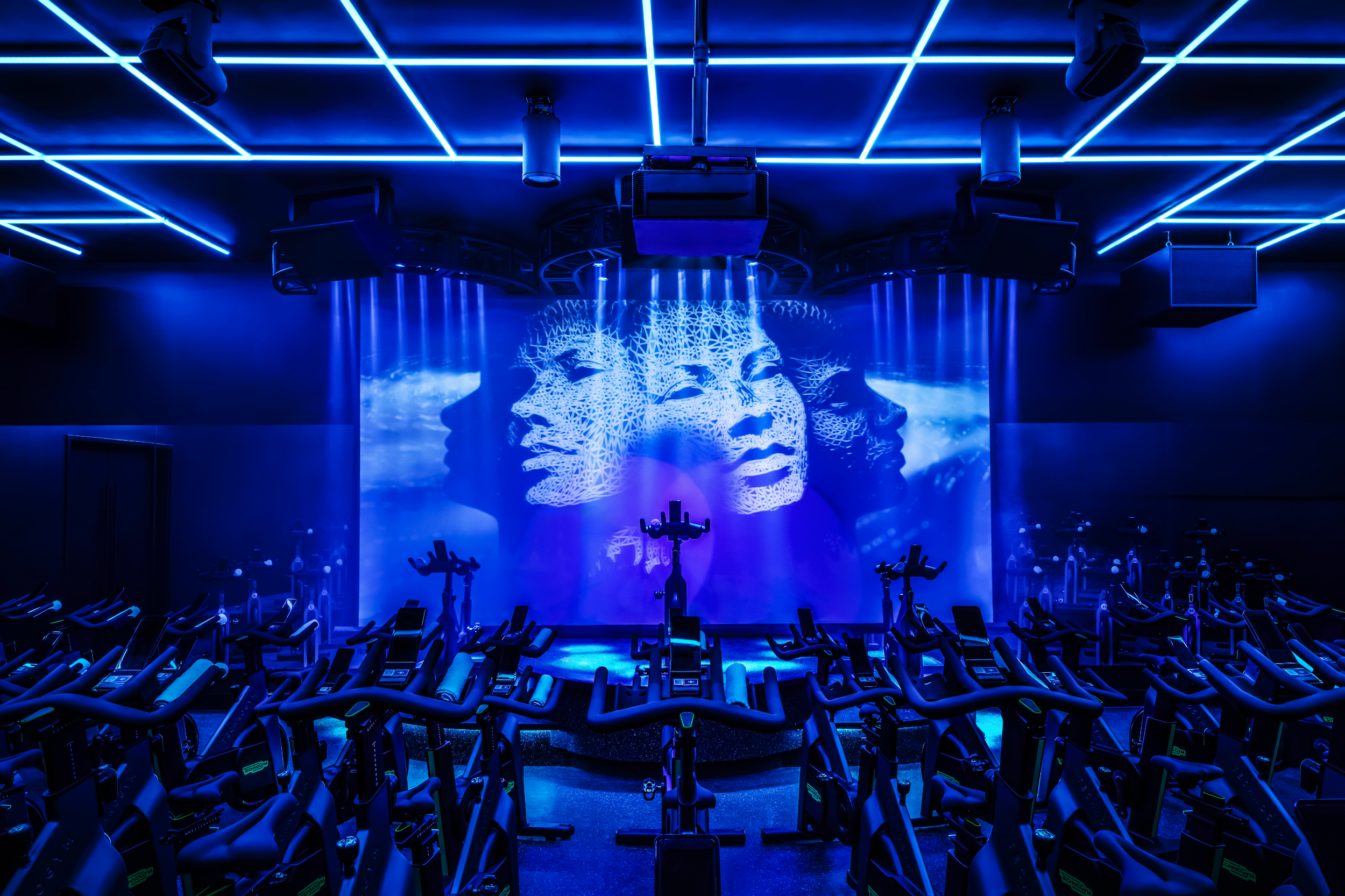Dark indoor cycling studio with purple and blue lighting, multiple stationary bikes arranged in rows, illuminated by stage lights and laser beams.