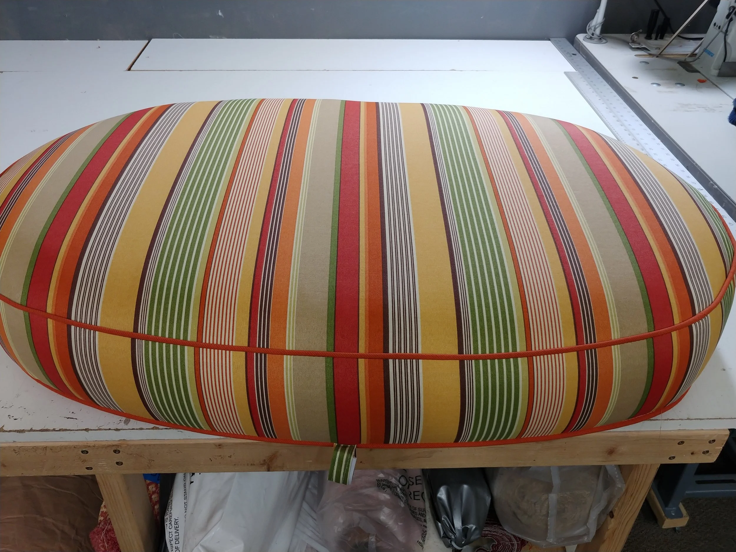 A colorful, striped oval cushion resting on a wooden table in a workshop. The cushion has various horizontal stripes in green, red, yellow, orange, beige, and black. part of a measuring ruler is visible on the right side of the table.