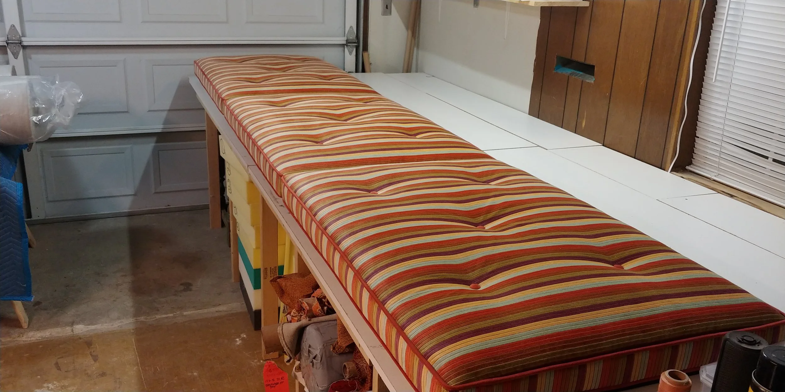 A long bed with a colorful striped mattress in a garage, with various boxes and items underneath.