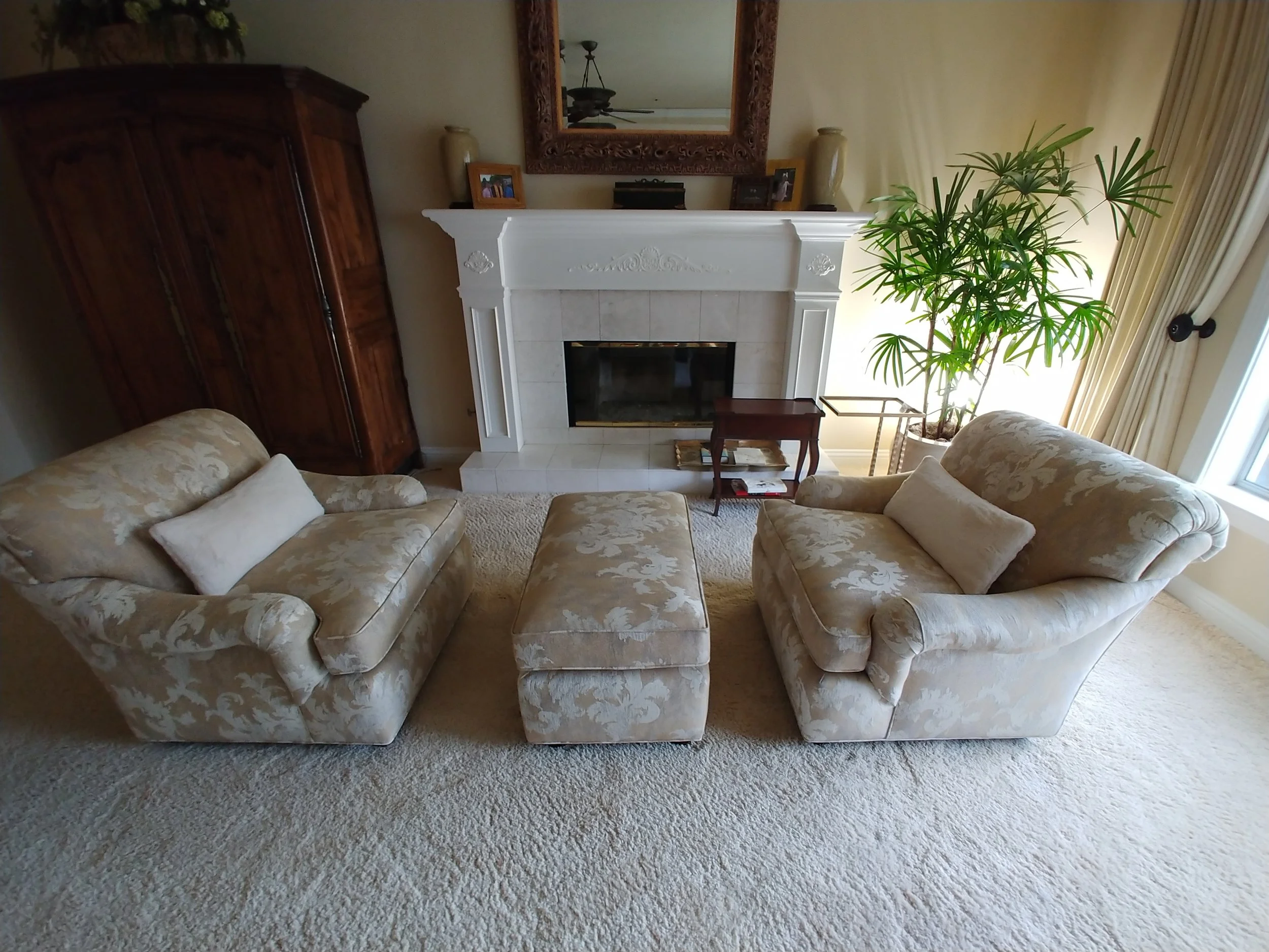 Living room with two floral-patterned sofas, a matching ottoman, a fireplace with a mirror above it, a large potted plant near a window, and various decorative items on the mantle and side tables.