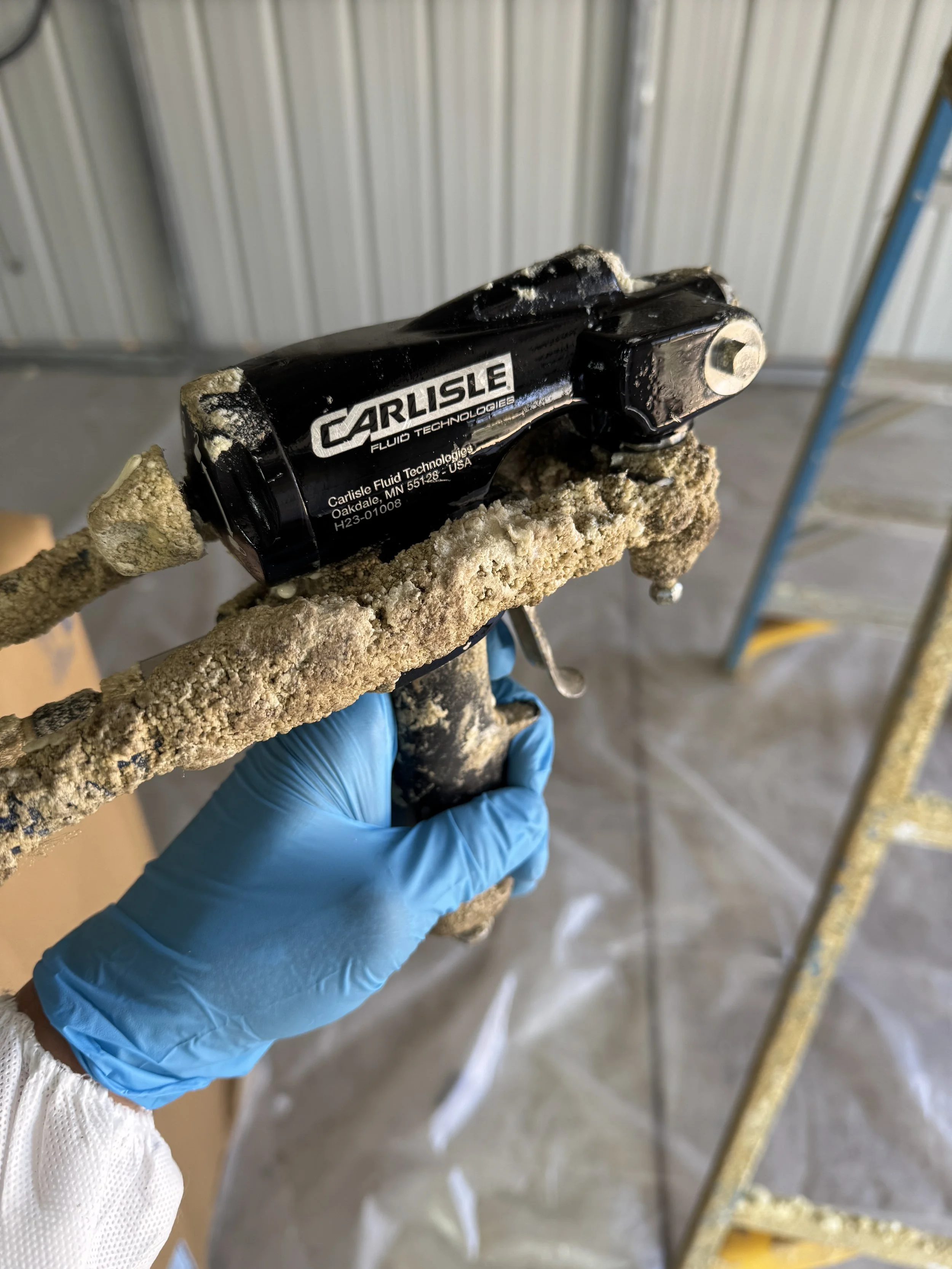 Person wearing a blue glove holding a spray foam gun  covered in foam in an industrial or construction setting.