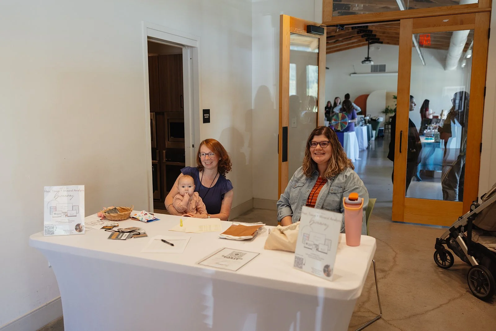 Two women sitting at a table with a baby, inside a room with a glass door leading to a larger event space with people, tables, and decorations.