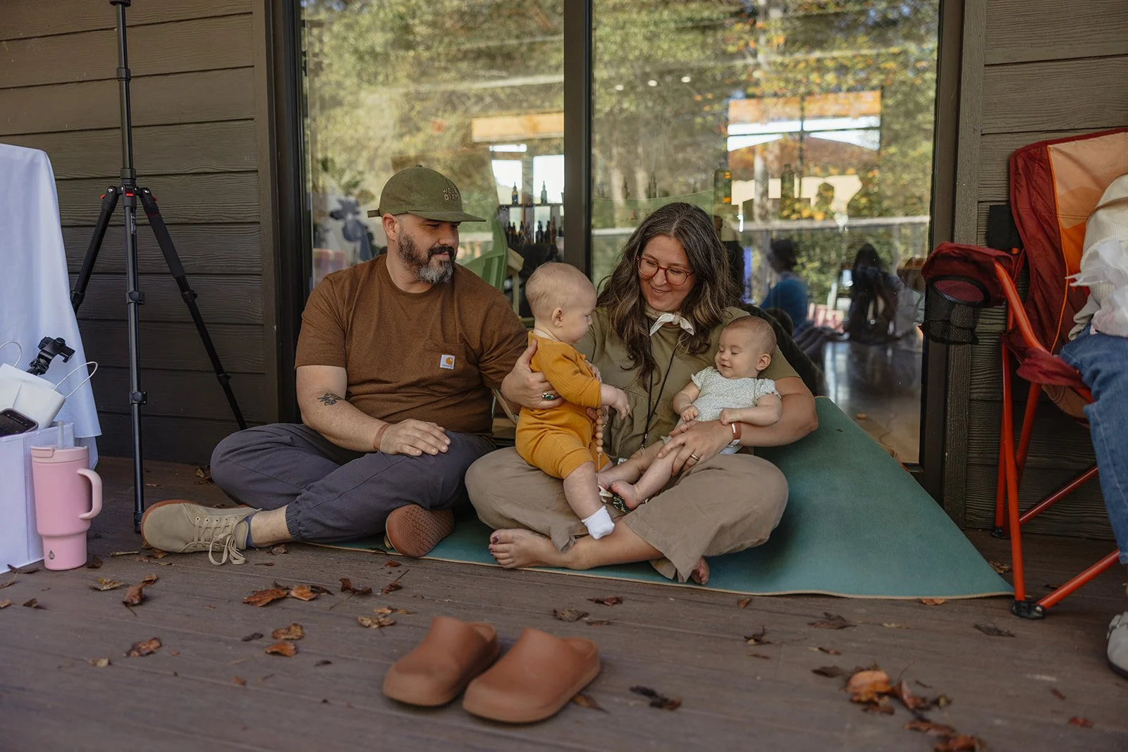 A family sitting on a green mat on a porch. The man, woman, and two babies are interacting, with one baby standing and the other baby sitting in the woman's lap. There are fallen leaves on the porch floor, and shoes placed nearby.