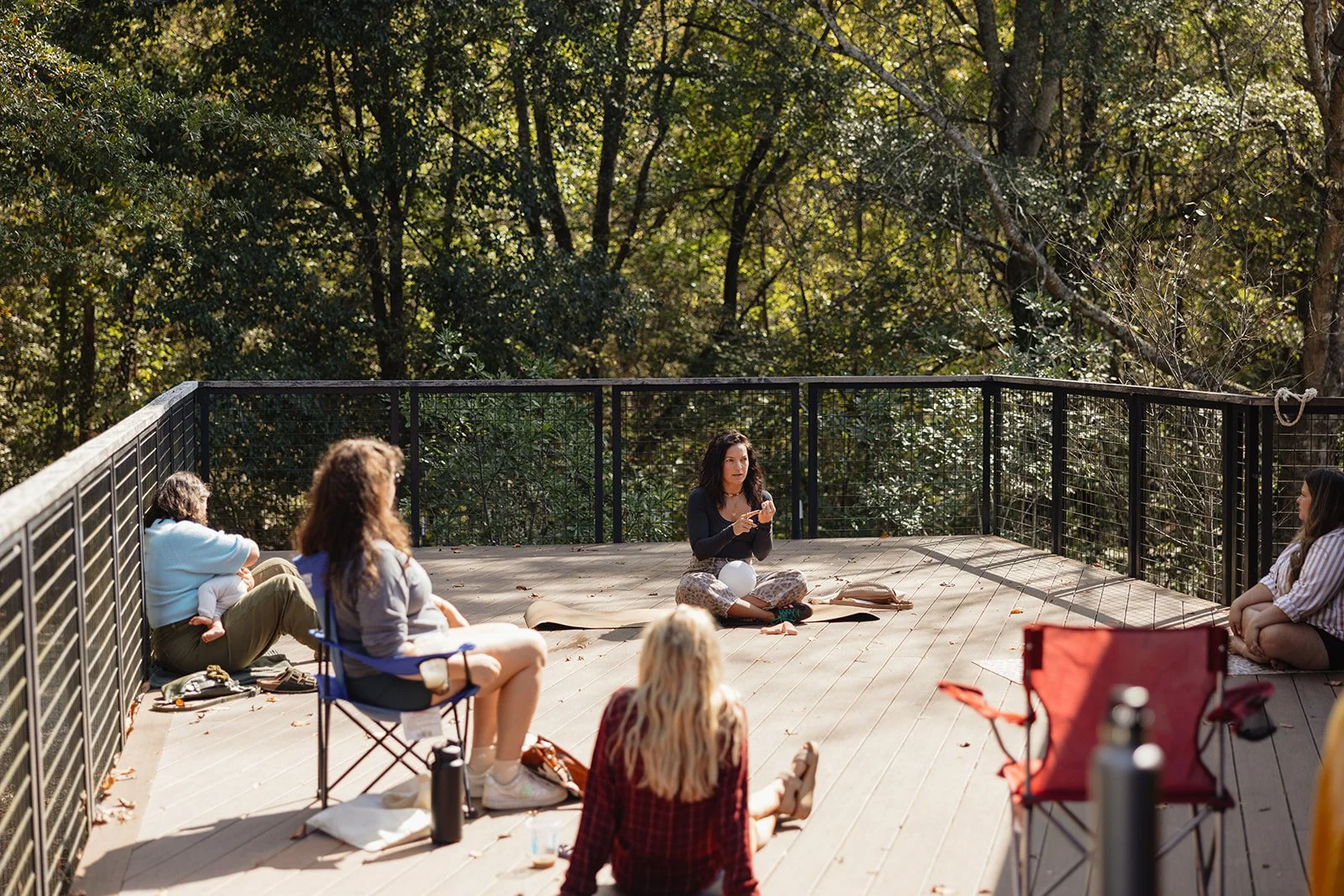 A group of women sitting on a wooden deck outdoors, practicing meditation or yoga, surrounded by trees.