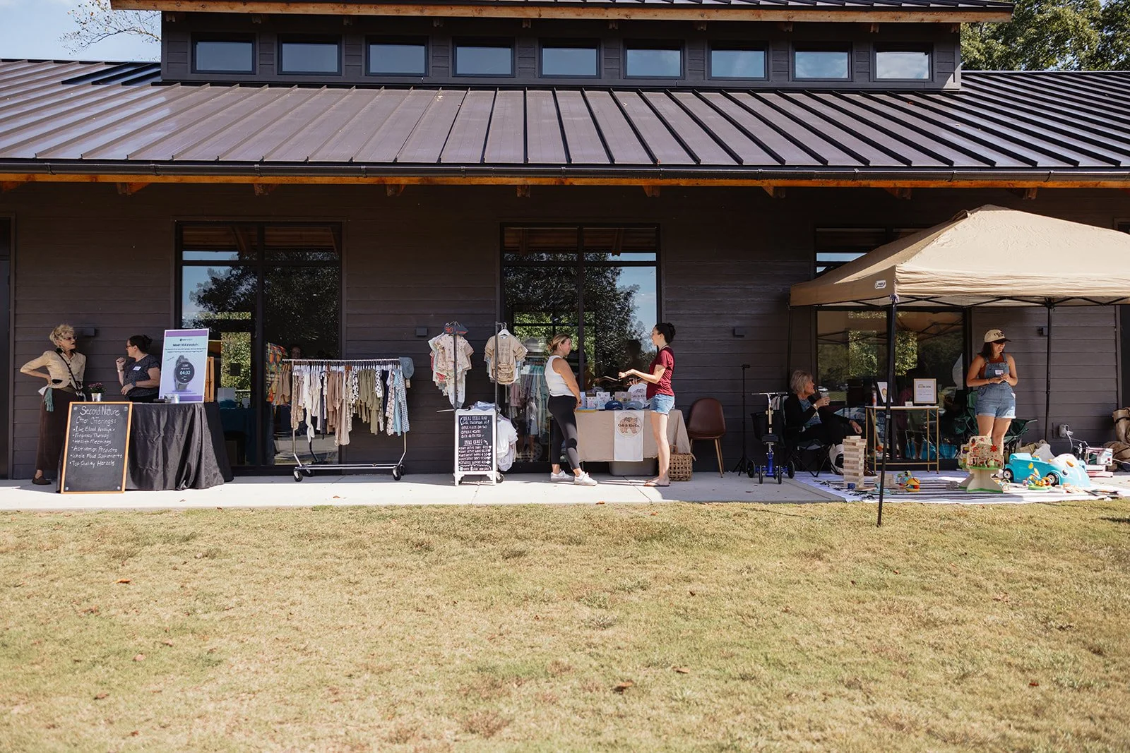 Outdoor flea market or craft sale with vendors and shoppers under a tent and in front of a dark wooden building.