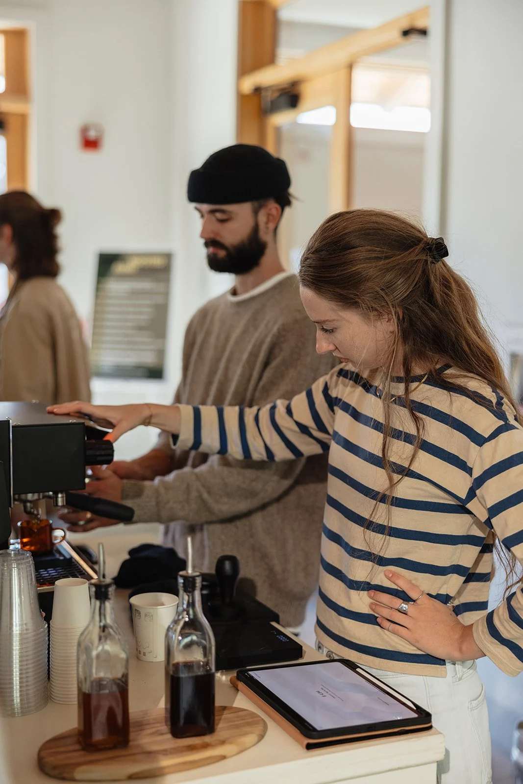 A woman in a striped shirt making coffee at a coffee shop, with a barista in the background.