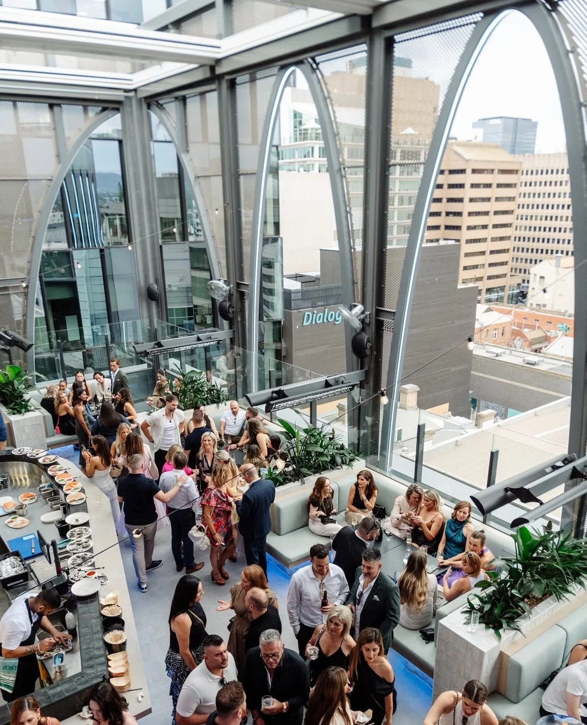 Indoor rooftop bar filled with people socializing, with large glass windows showing city buildings outside.
