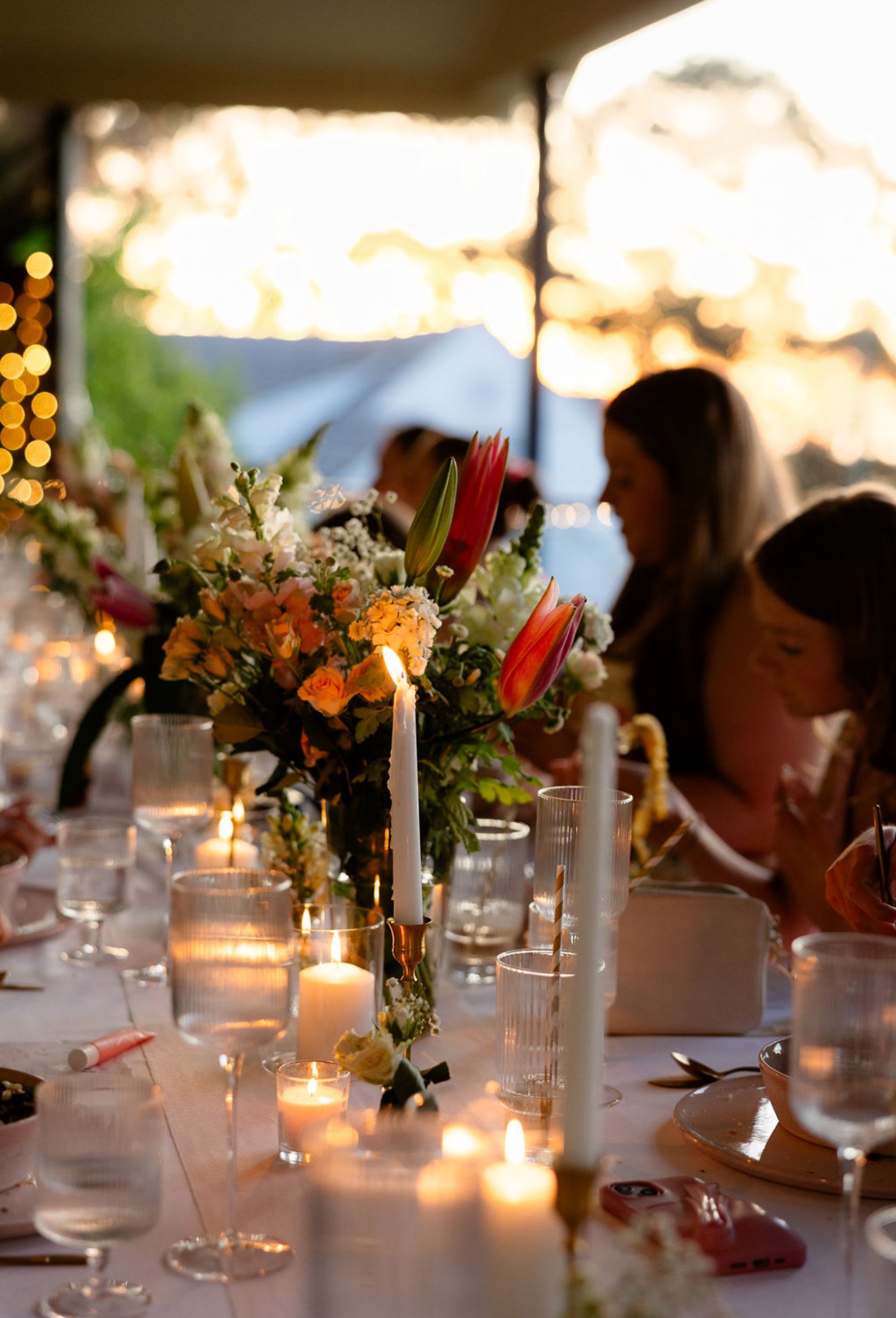 A dinner table decorated with candles and a colorful bouquet of flowers, with people seated and a sunset visible in the background.