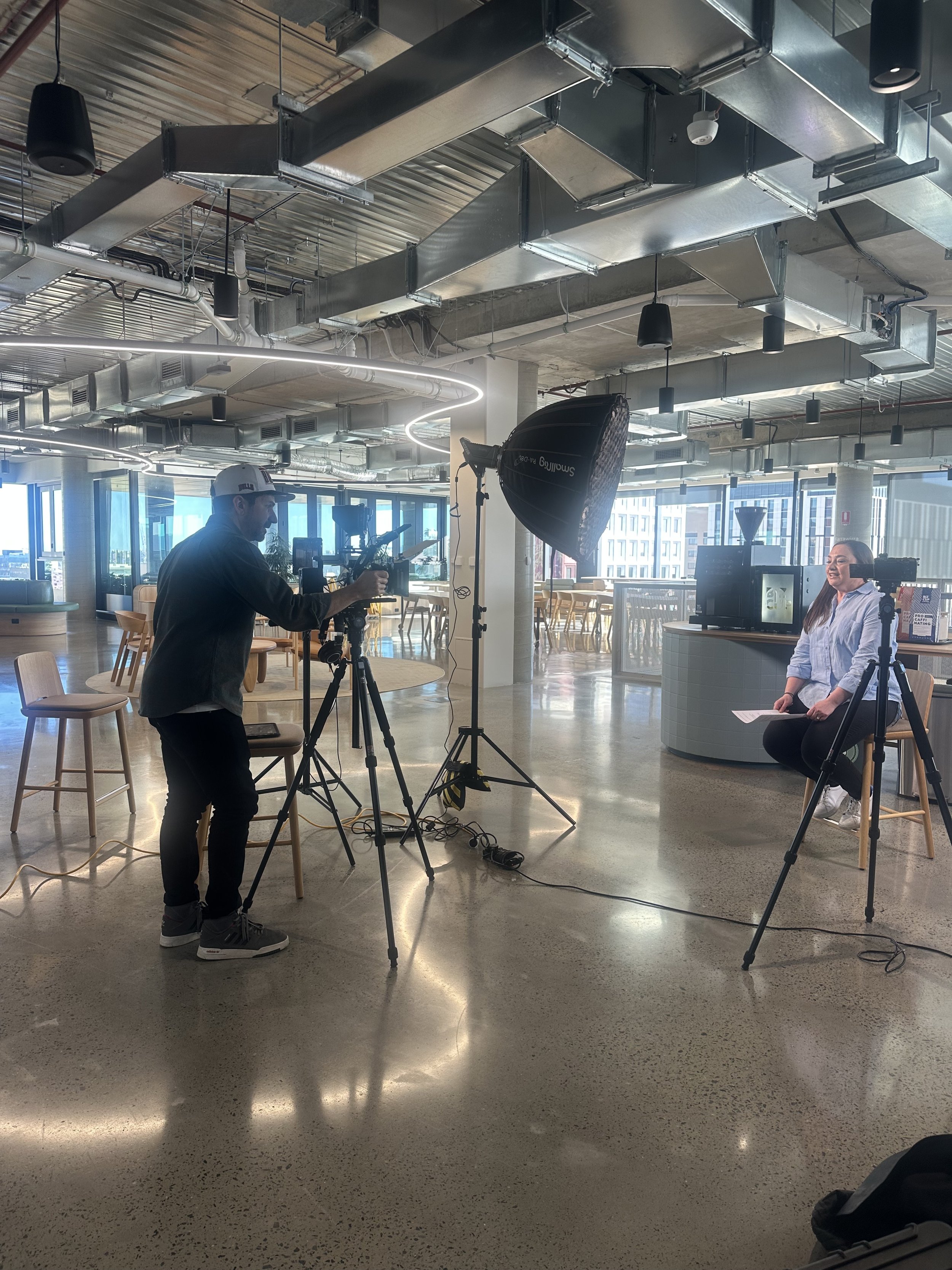 A woman being filmed in a studio with professional lighting and camera equipment, in a modern office space with large windows.