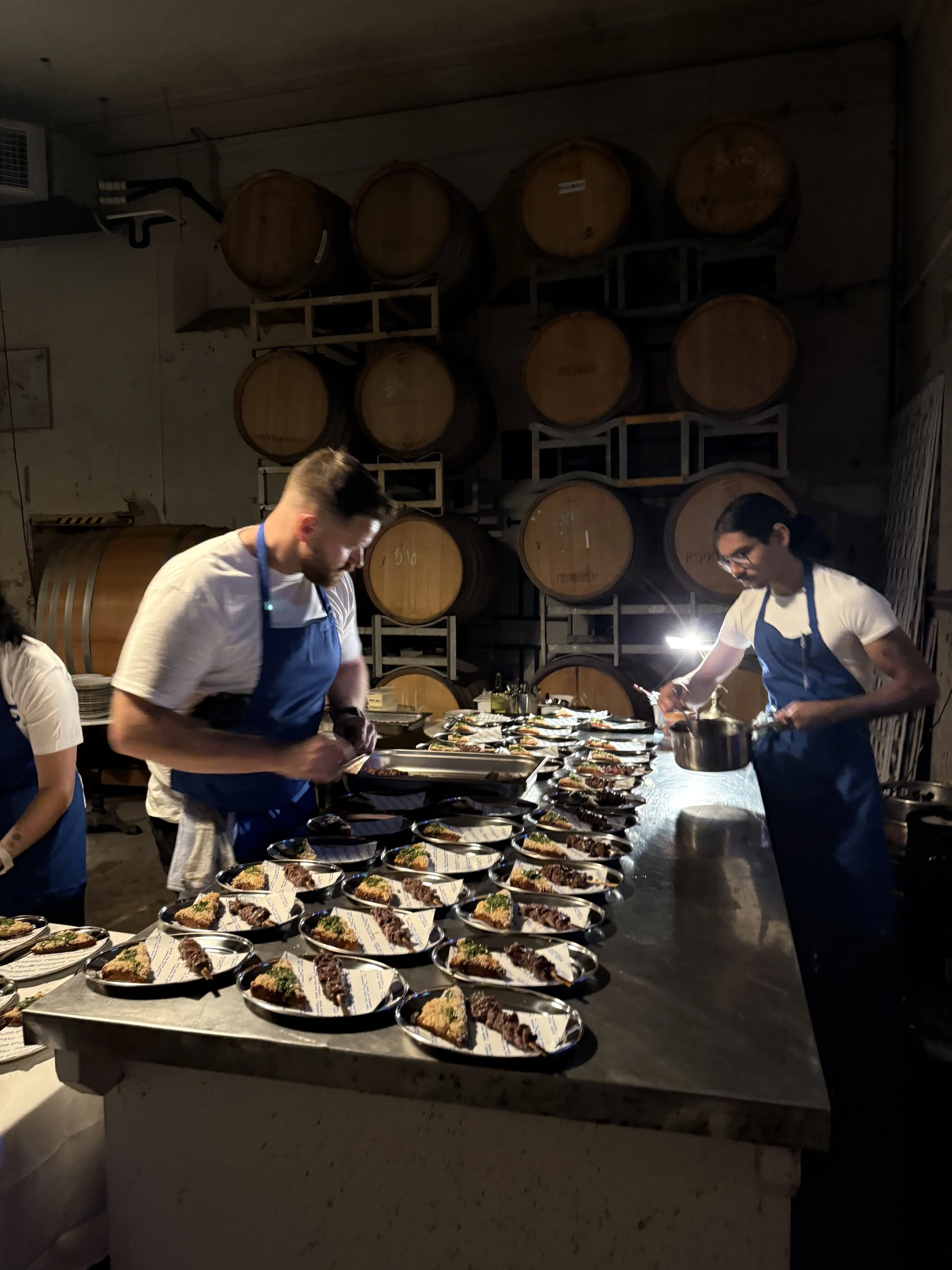 Two chefs preparing a large number of plated dishes in a dimly lit kitchen with wine barrels stacked in the background.