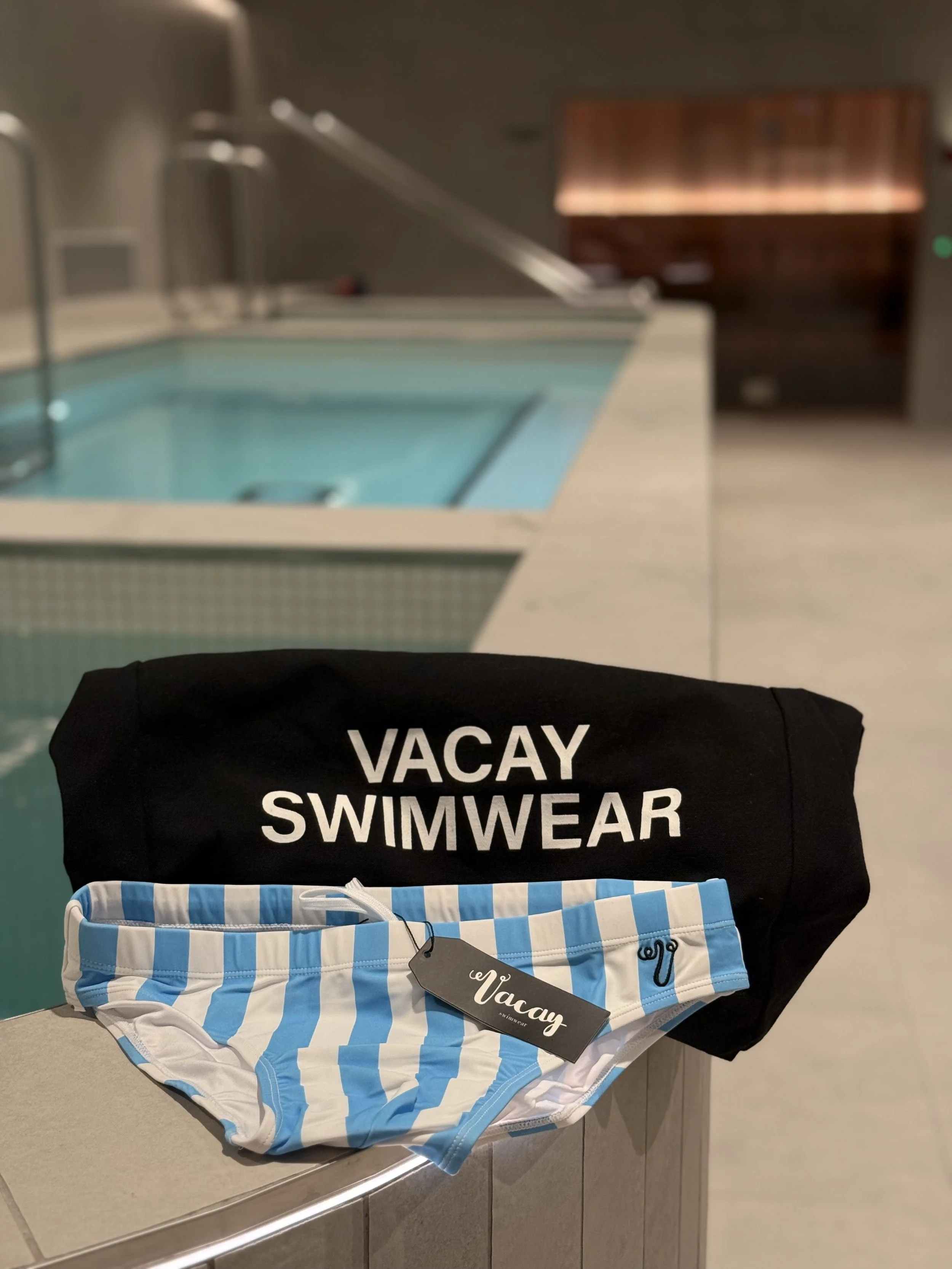 Blue and white striped Swimmmear briefs displayed in a retail store, with a black 'VACAY' swimwear bag behind them.