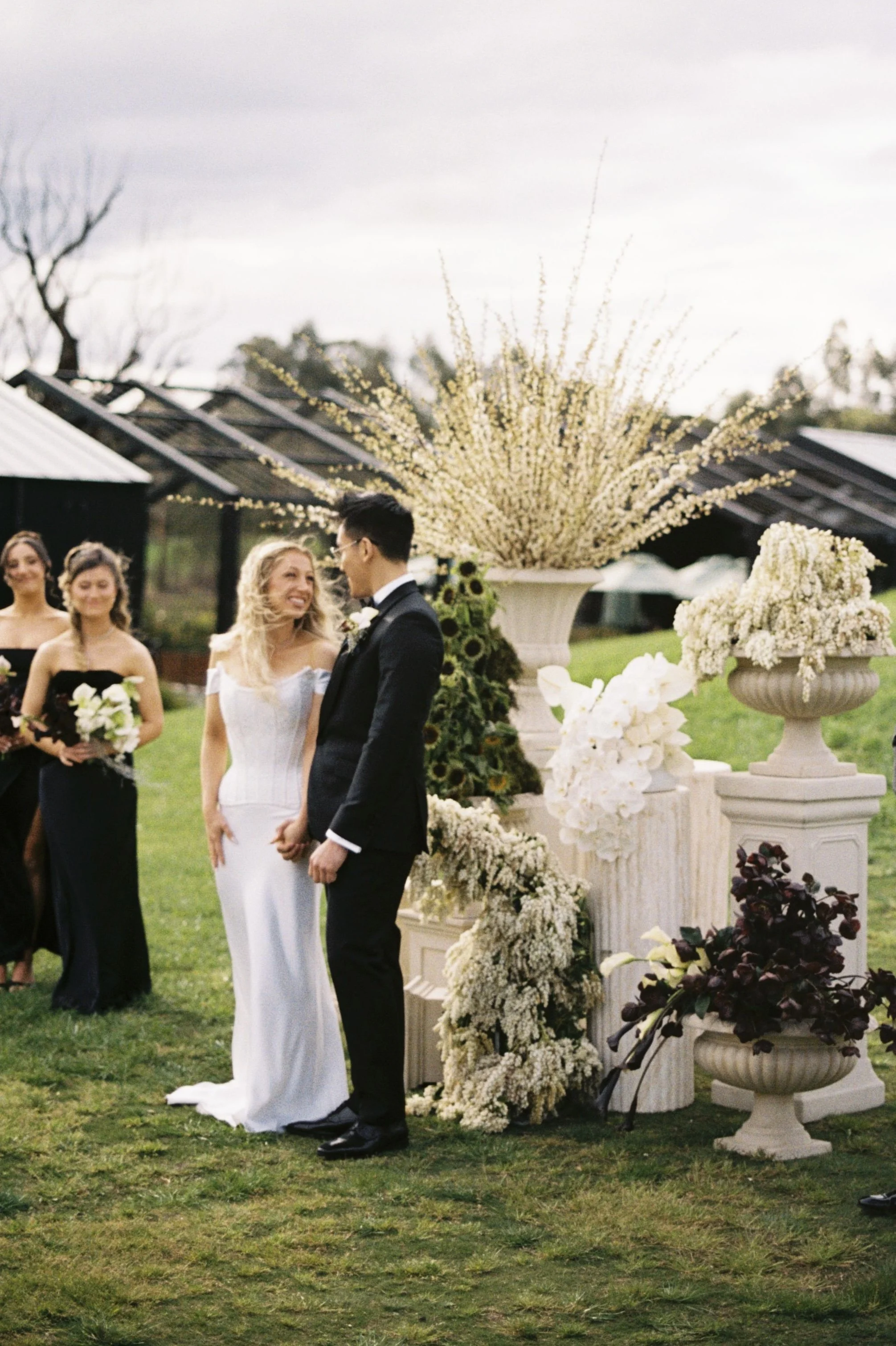A bride and groom holding hands during their outdoor wedding ceremony, surrounded by bridesmaids and large floral arrangements.