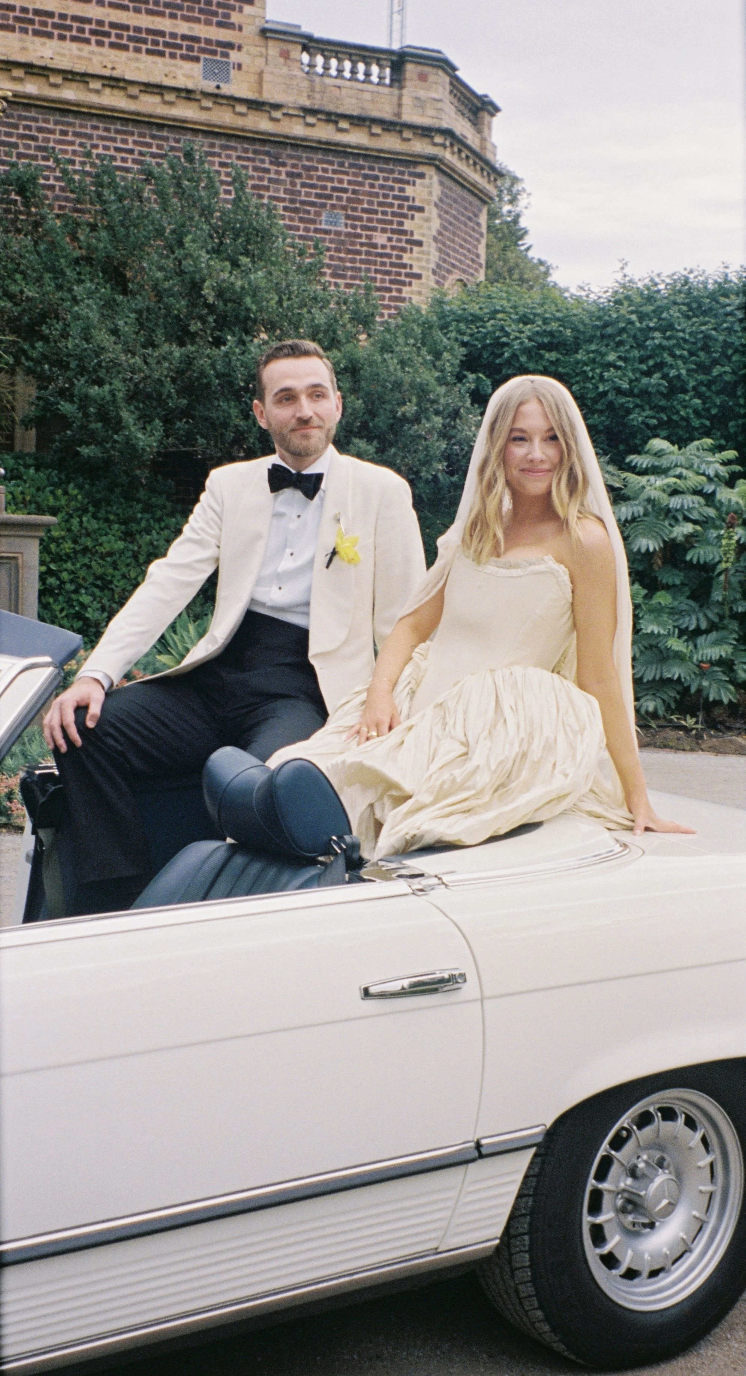 A man and woman dressed in wedding attire sitting in the back of a classic white convertible car, with a lush garden and brick building in the background.