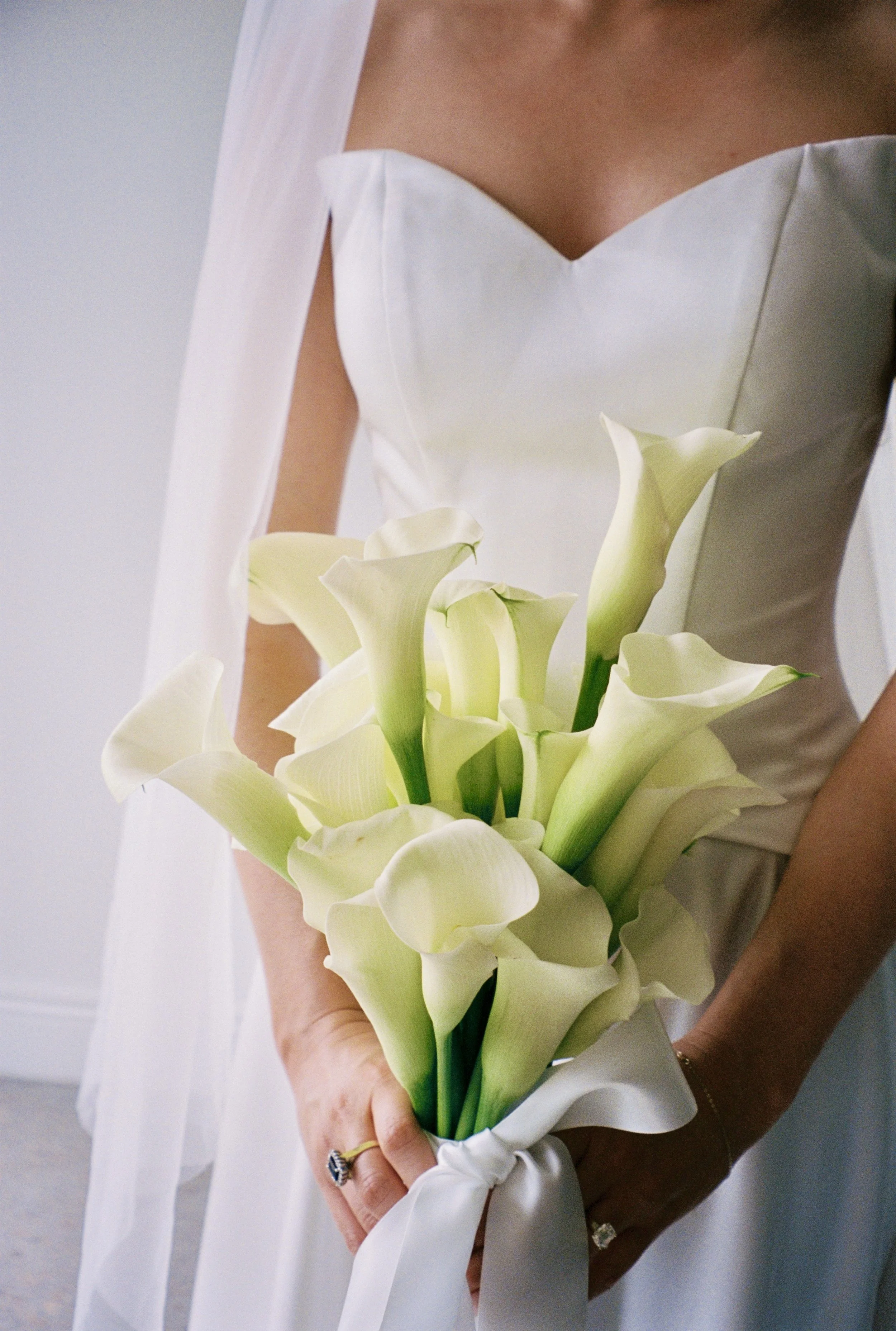 A woman wearing a white gown holding a bouquet of white calla lilies with a white ribbon.