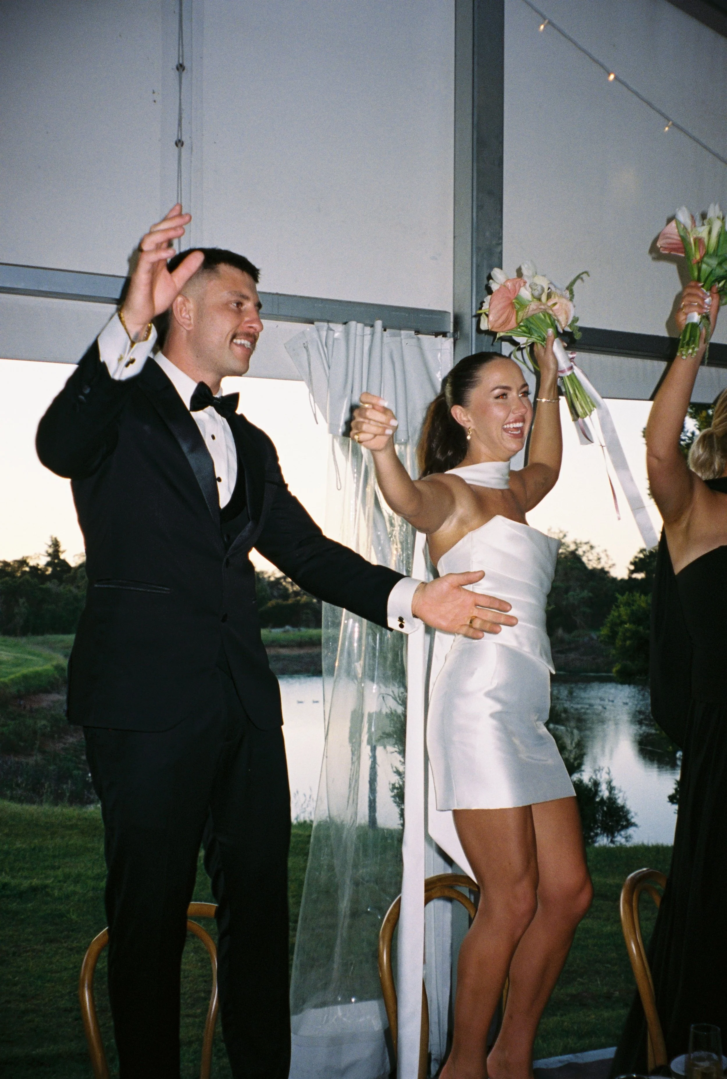 Bride and groom celebrating with raised arms and bouquets during wedding reception near window overlooking river and trees.