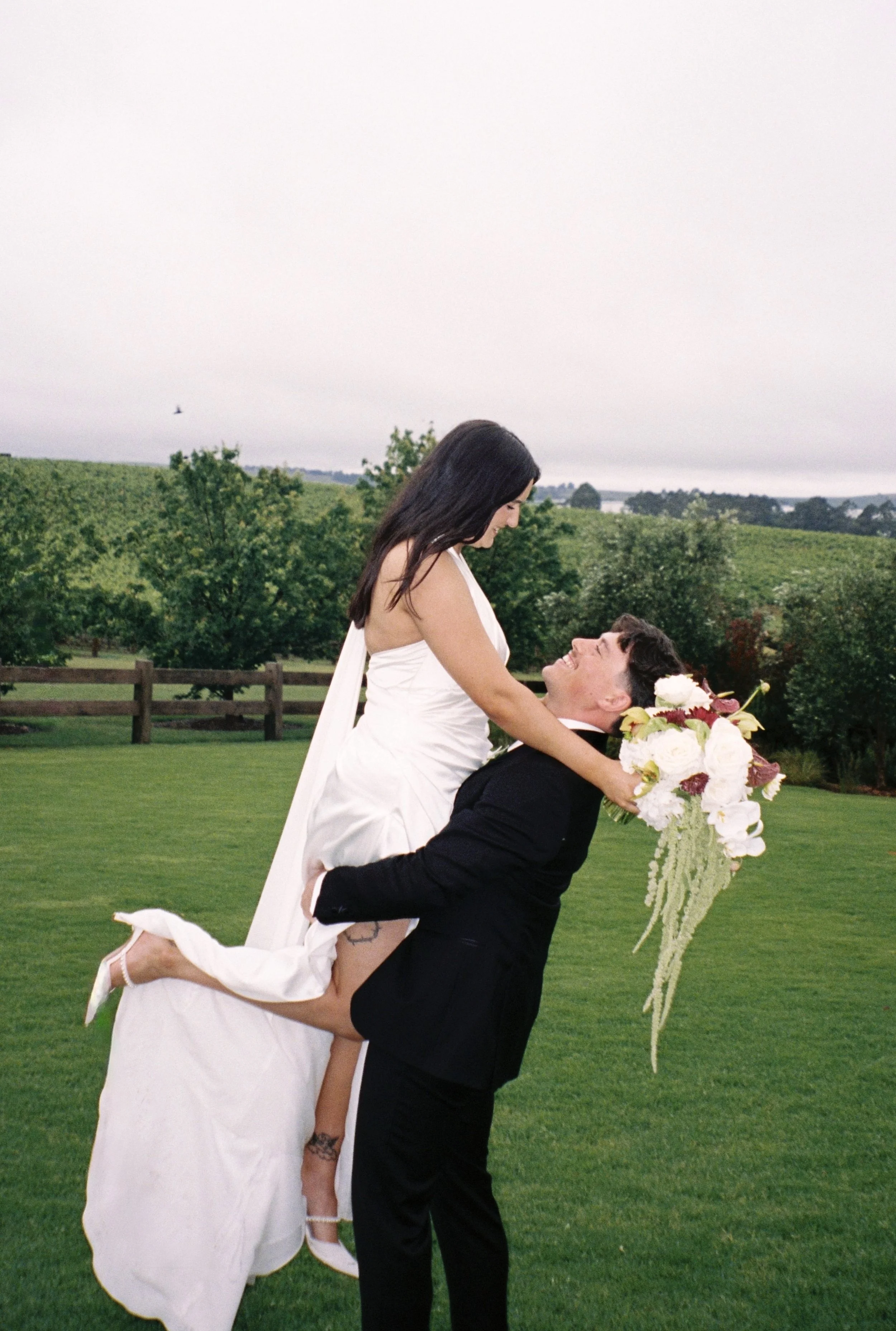 A bride and groom outside on a green lawn, with the groom lifting the bride and holding a large bouquet of flowers, with trees and a fence in the background.