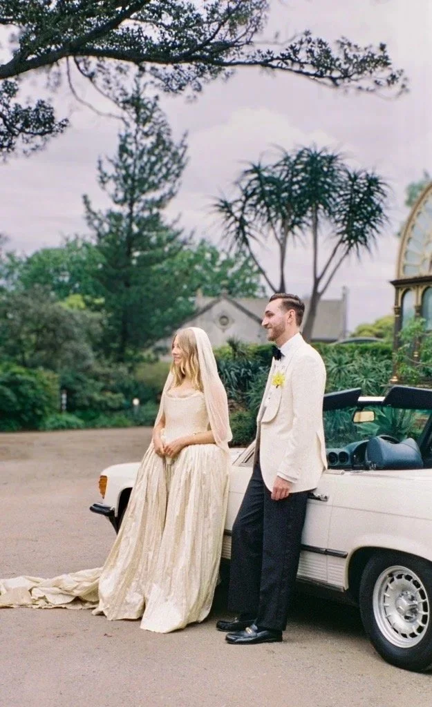 A bride and groom in formal attire standing beside a classic white convertible car outdoors, surrounded by trees and green foliage.