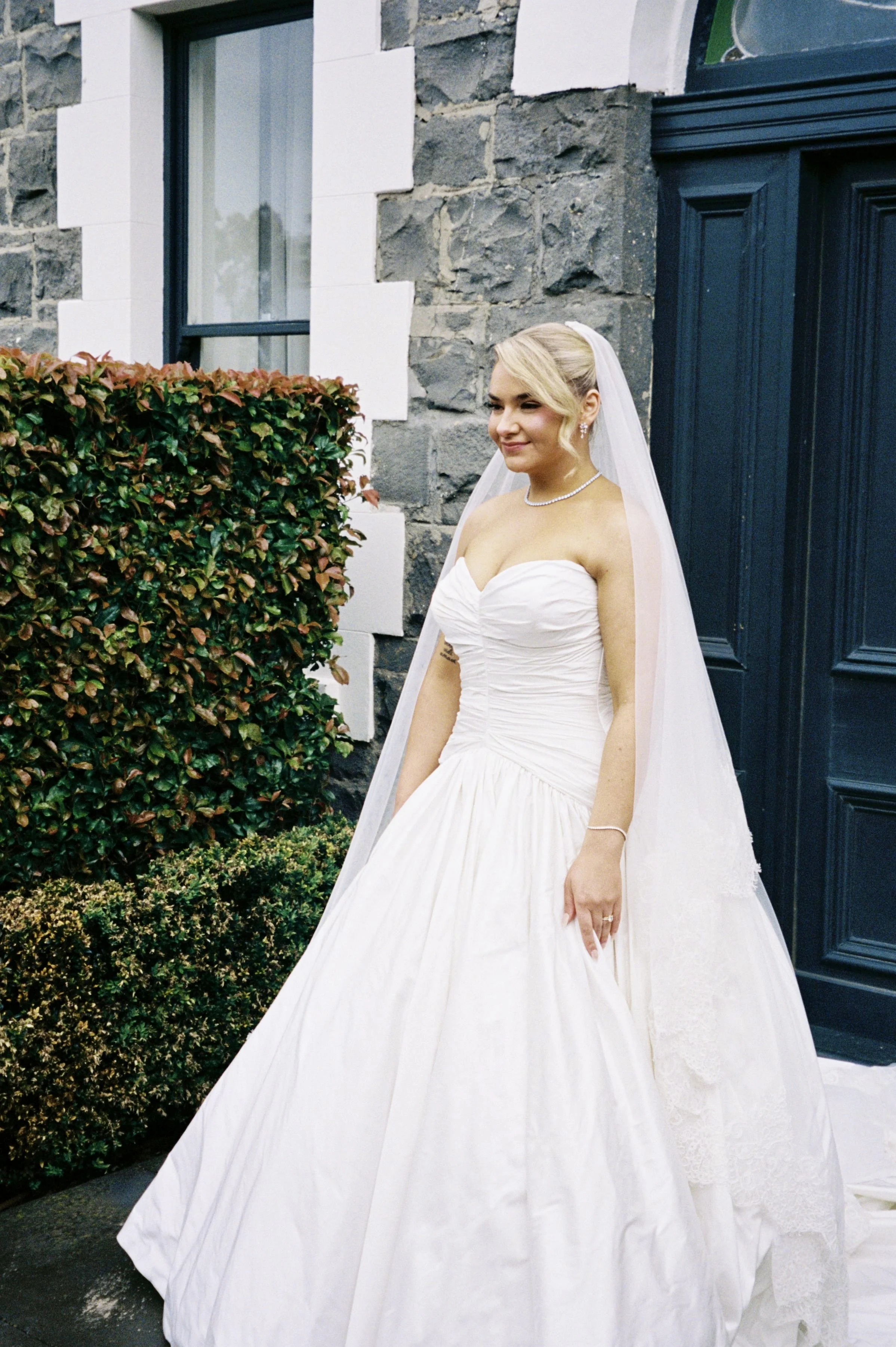 A bride in a strapless white wedding gown with a veil standing outside near a stone building and greenery.