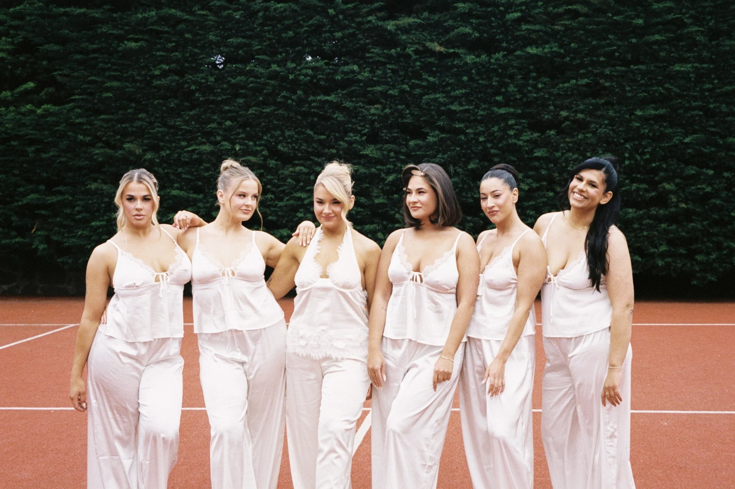 Six women in matching white pajamas standing on a tennis court, with large green bushes in the background.