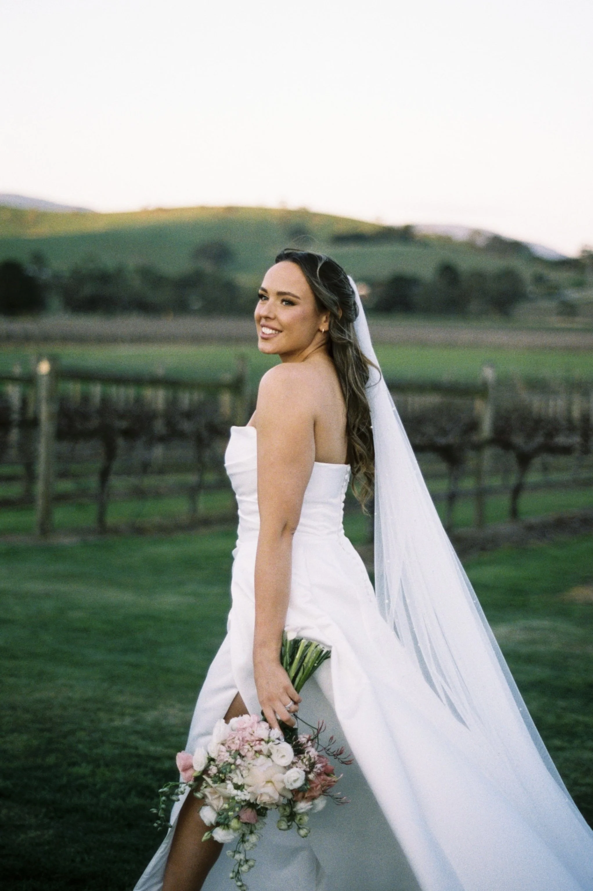 A bride in a strapless white wedding gown, holding a bouquet of pink and white flowers, standing outdoors on a green lawn with rolling hills and vineyard in the background.
