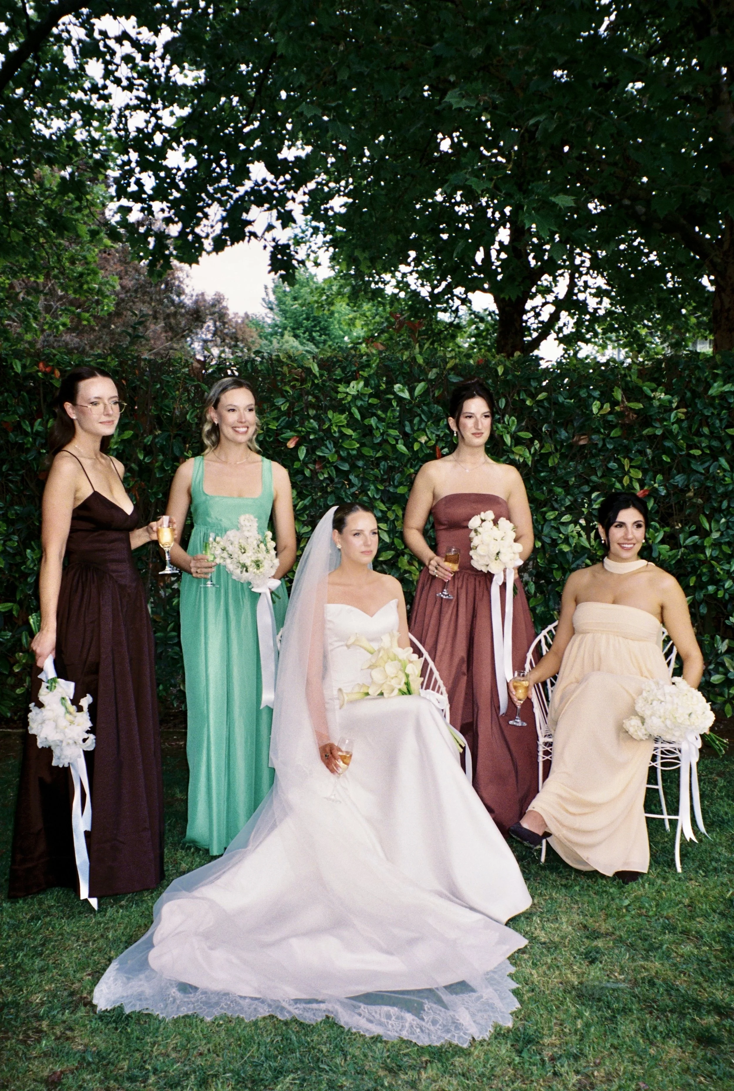 A bride in a wedding dress with a veil and gloves sitting on a white chair, surrounded by five women in colorful dresses, all holding bouquets and glasses of champagne, outdoors in front of green shrubs.