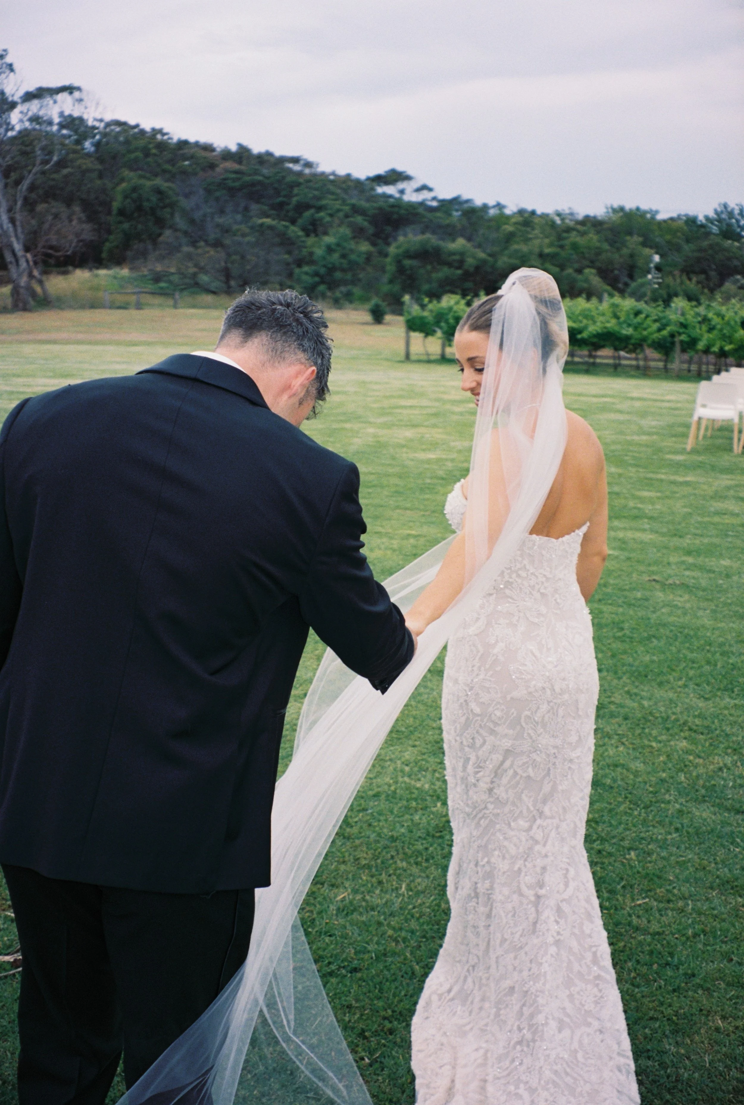 A groom in a black suit and a bride in a white strapless wedding dress with a long veil are holding hands outdoors on a grassy field, with trees and chairs in the background.