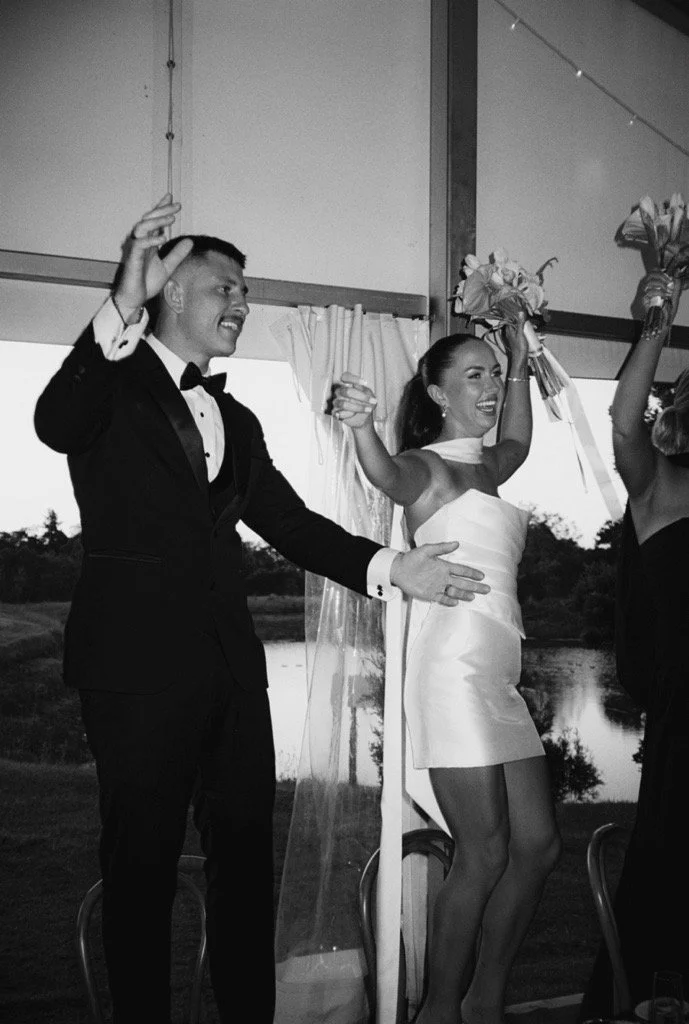 A couple celebrating at a wedding reception, smiling and raising their hands, the woman holding a bouquet of flowers, in front of a window with a scenic view.