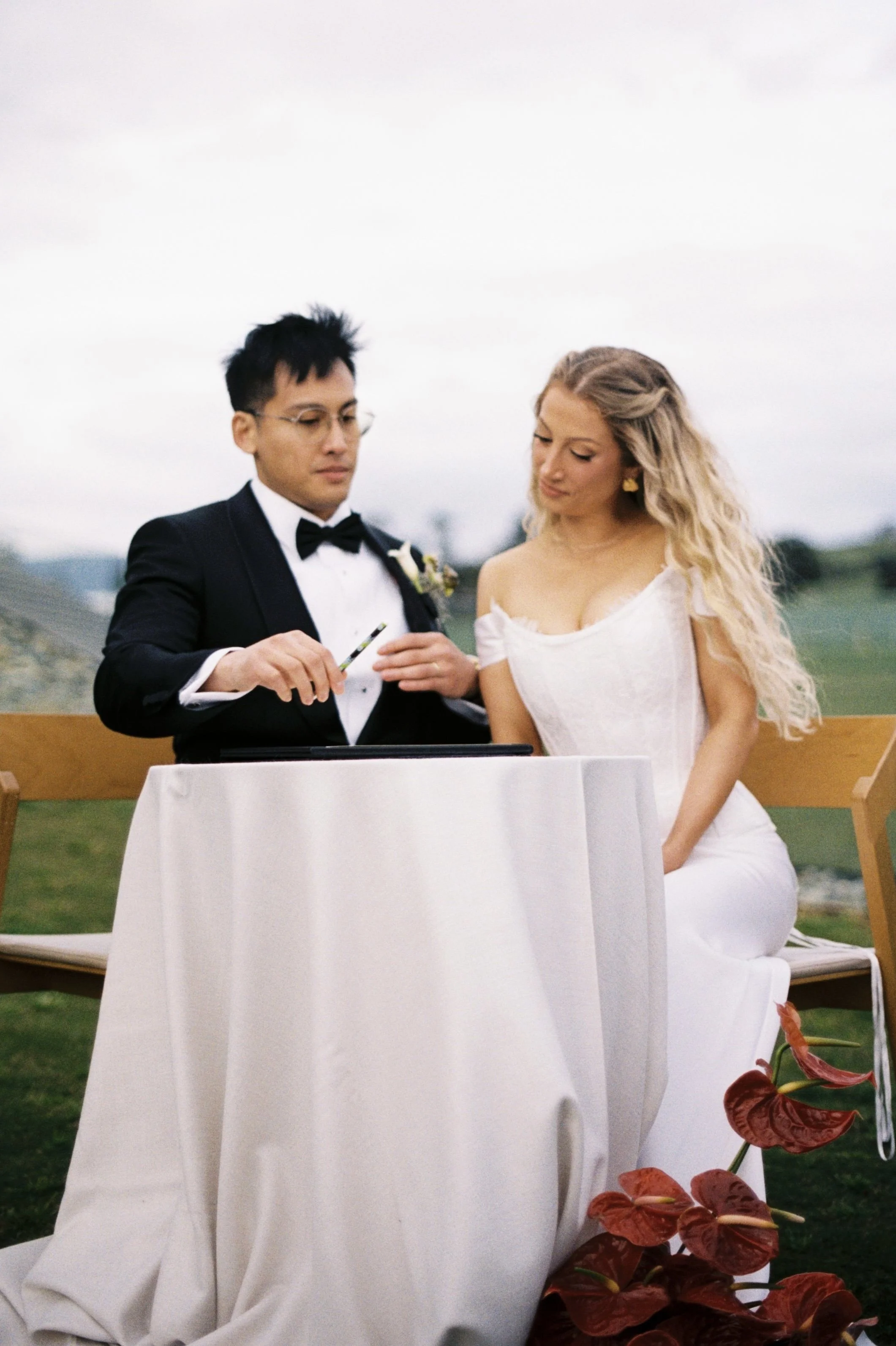 A couple getting married outdoors, with the groom in a black tuxedo and the bride in a white dress, standing at a small table covered with a white cloth.