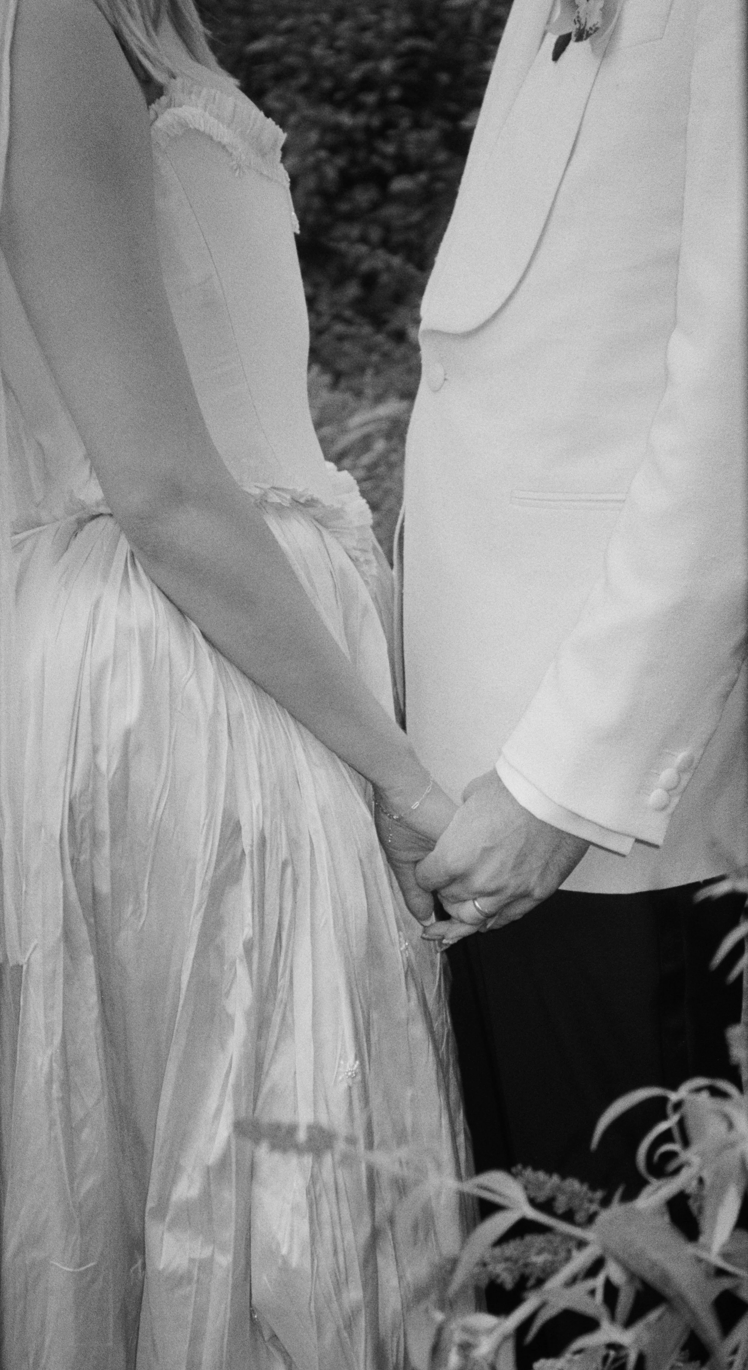 Close-up of a bride and groom holding hands during a wedding, with the bride wearing a wedding dress and the groom in a suit, in black and white.