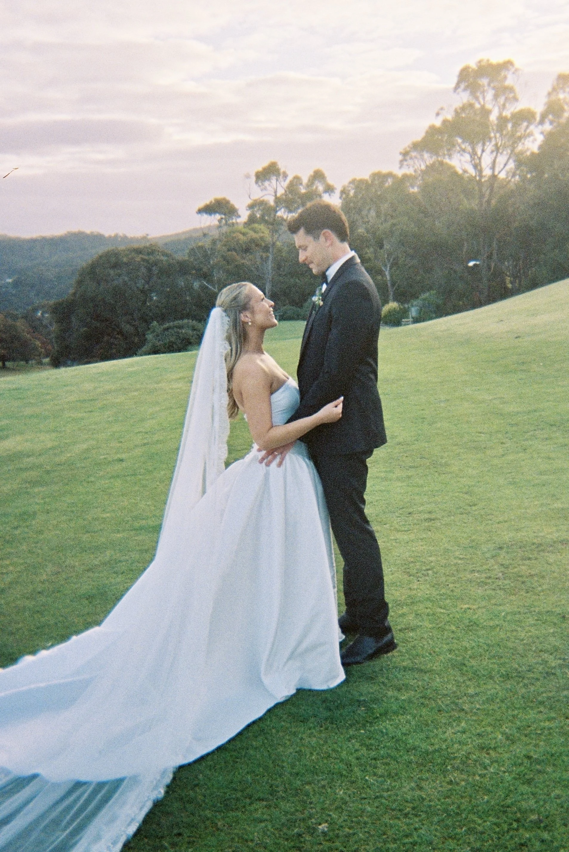 A bride and groom standing on a grassy field, gazing into each other's eyes during sunset, with trees and hills in the background.