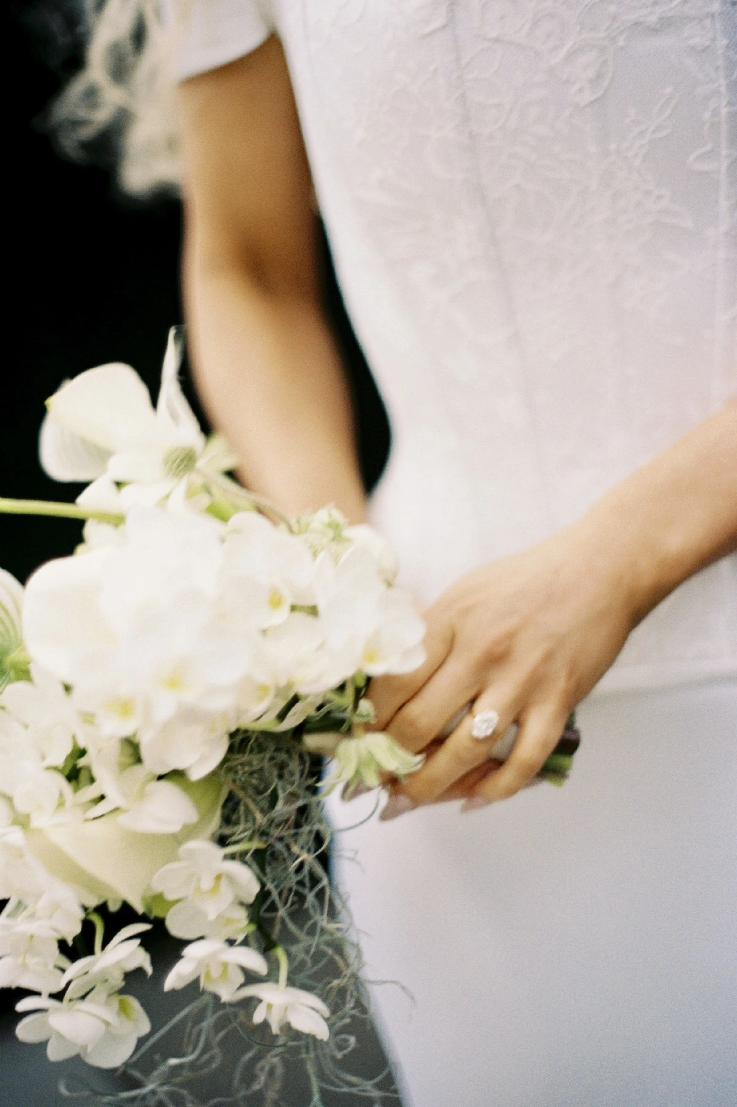Close-up of a person holding a bouquet of white flowers with their hand, wearing a ring, and dressed in white and black.