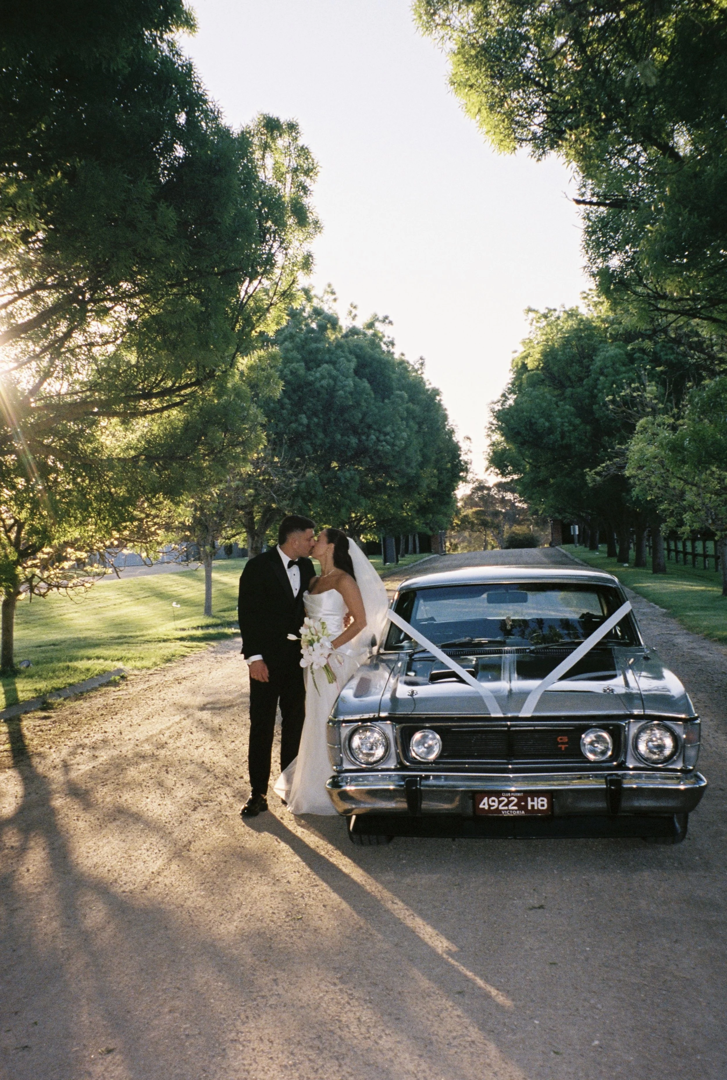 A newlywed couple dressed in wedding attire kissing beside a classic black car decorated with white ribbons, parked on a tree-lined road during sunset.