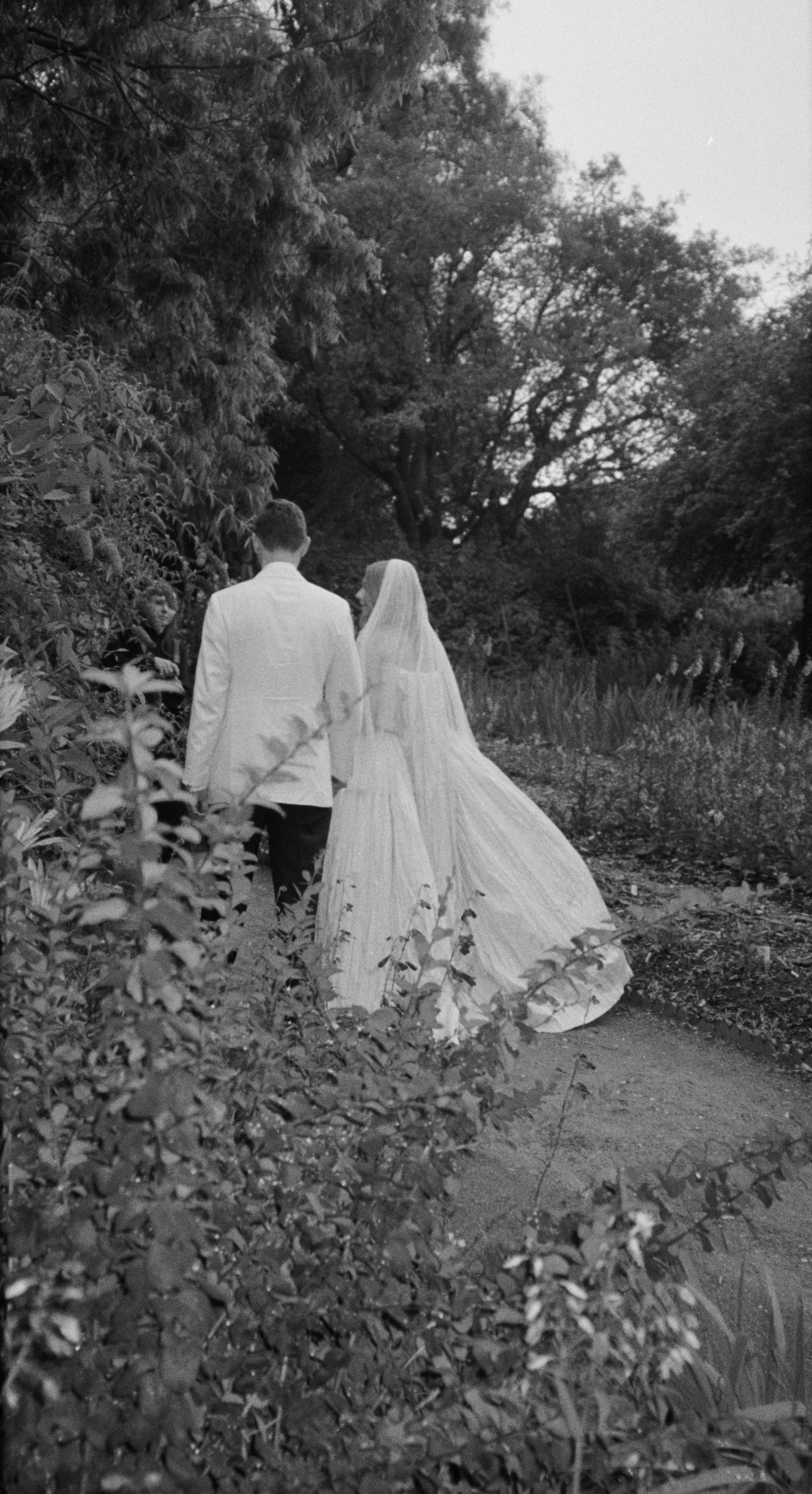 A black-and-white photo of a bride and groom walking along a wooded path during their wedding, surrounded by trees and bushes.