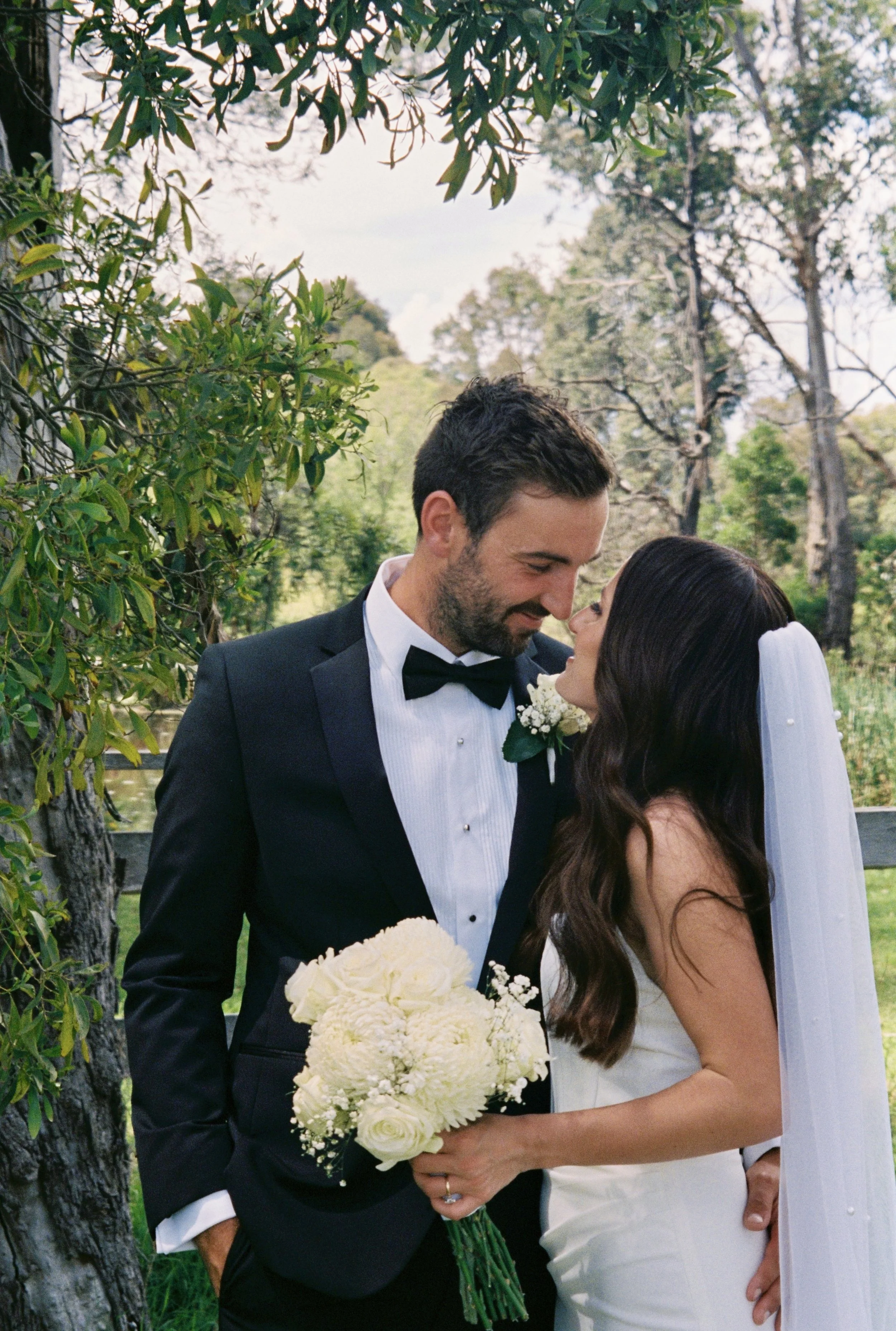 A bride and groom in wedding attire sharing a close, intimate moment outdoors surrounded by trees.