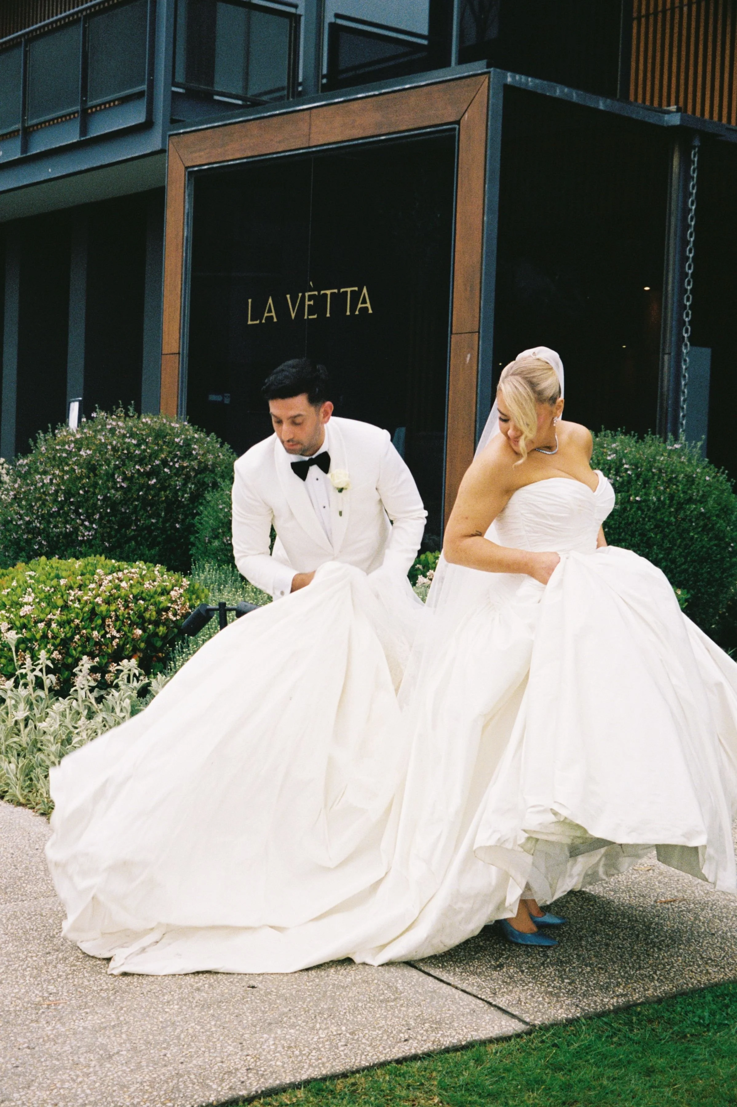 A bride and groom in wedding attire pose outside a building with a black sign that reads 'LA VÉTTA'. The bride is in a strapless white wedding gown, and the groom is in a white tuxedo with a black bow tie, both adjusting their dresses.