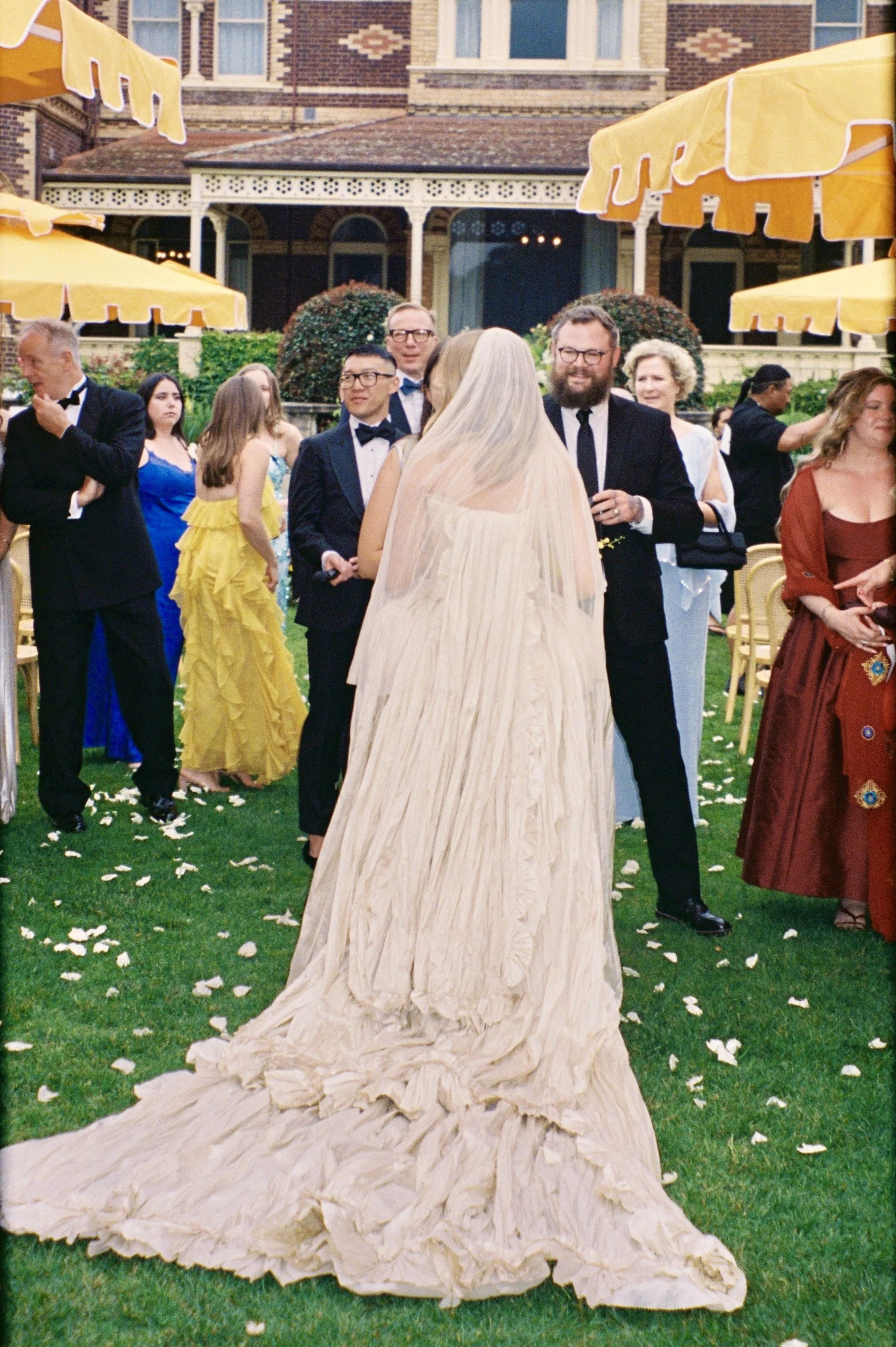 Bride in a cream-colored wedding gown and veil standing outdoors on a grassy area, surrounded by wedding guests dressed in formal attire, with some in evening gowns and tuxedos, at a garden wedding reception with yellow umbrellas and a large house in