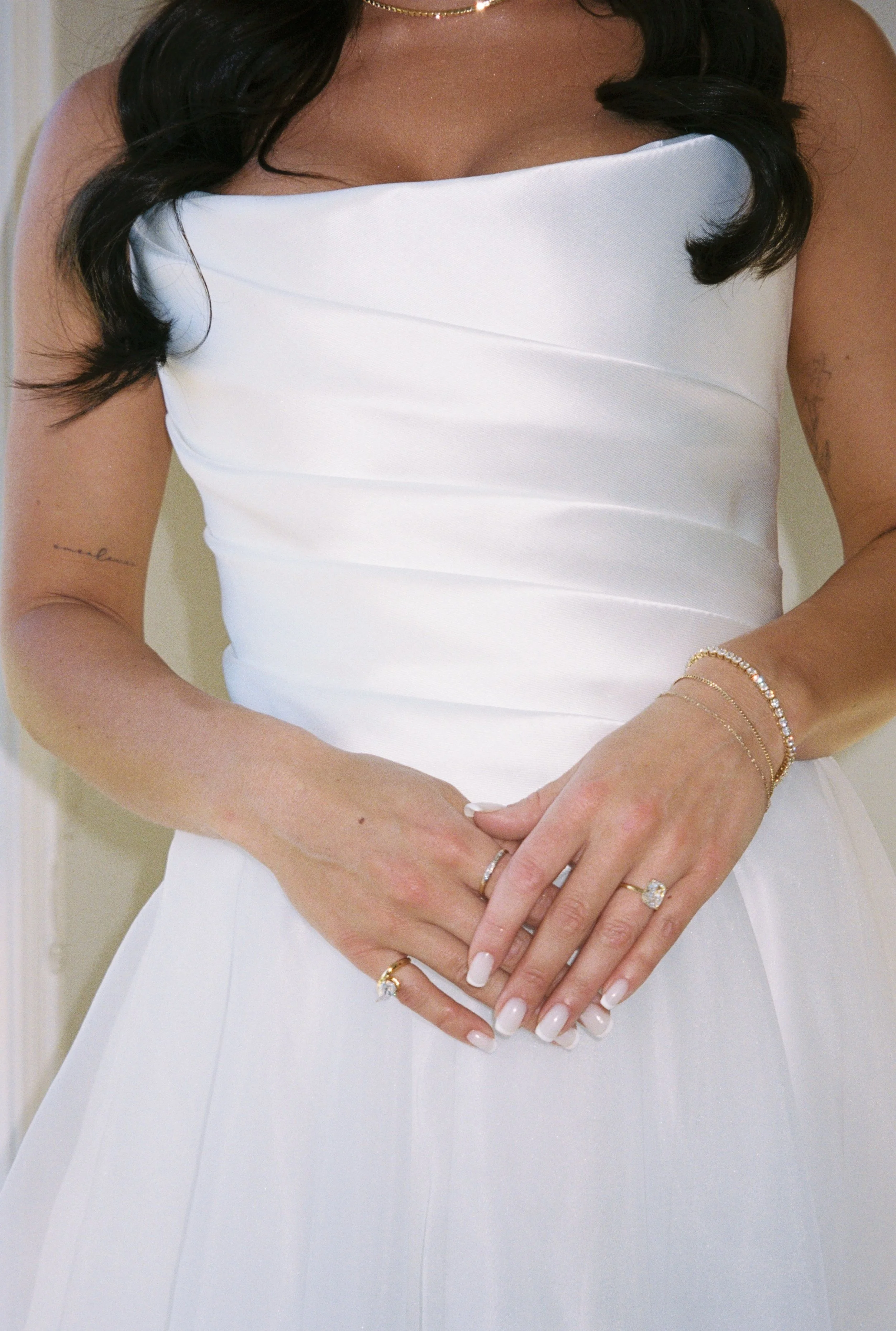 Close-up of a bride in a white strapless wedding dress, with dark hair and jewelry, her hands clasped together showing rings and bracelets.