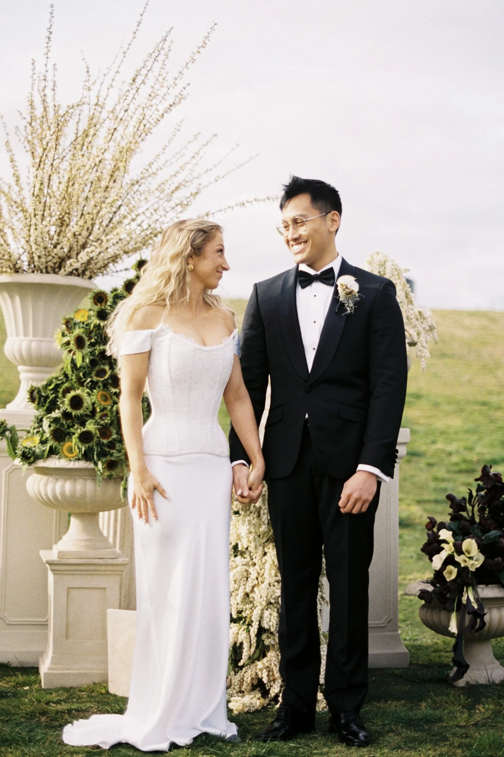 A bride and groom holding hands and smiling during their outdoor wedding ceremony, with floral arrangements and vases of flowers in the background.