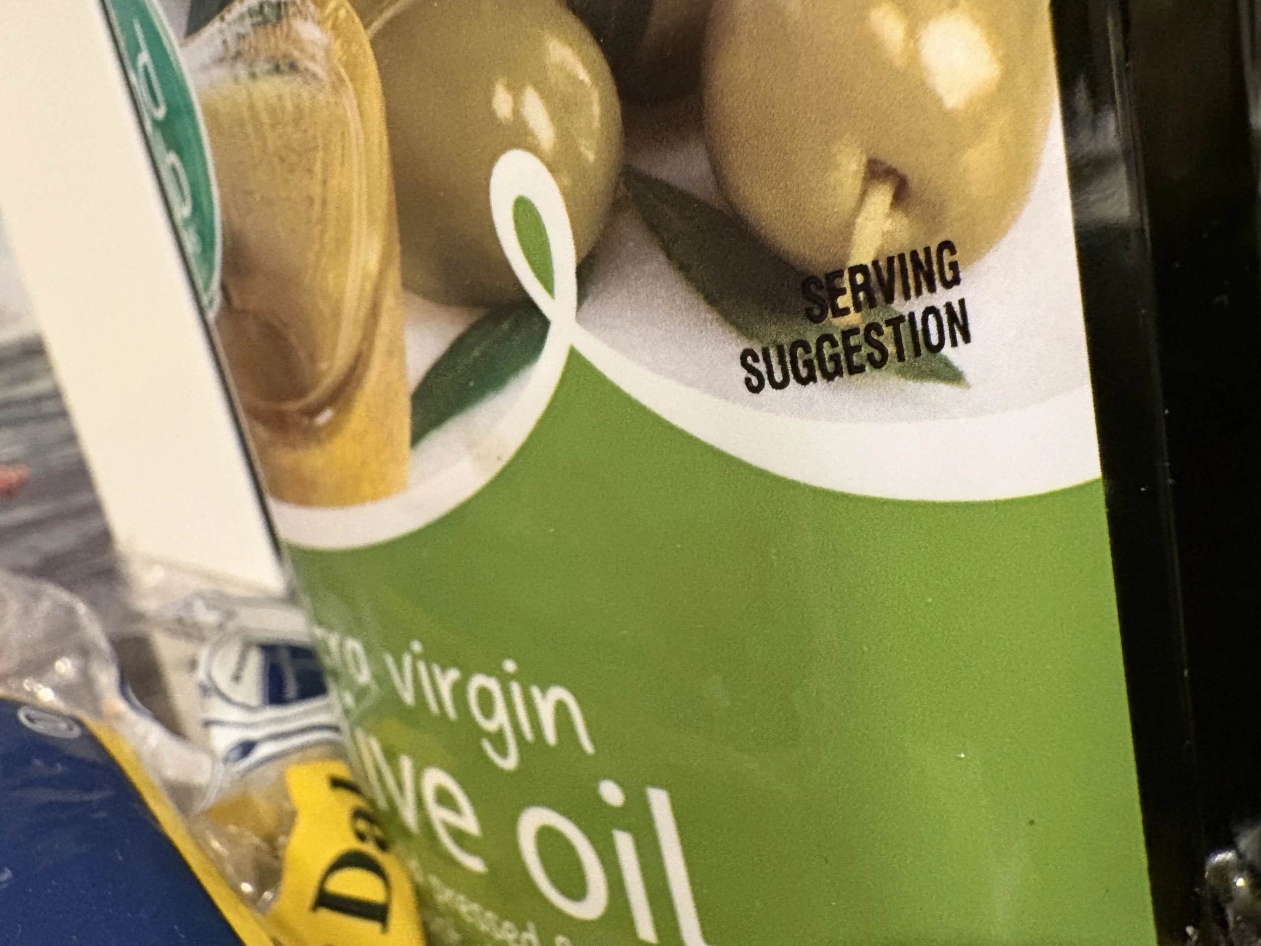 Close-up of a green and white package of avocado virgin oil with a serving suggestion printed on it. Part of a yellow and blue bag and other groceries are visible nearby.