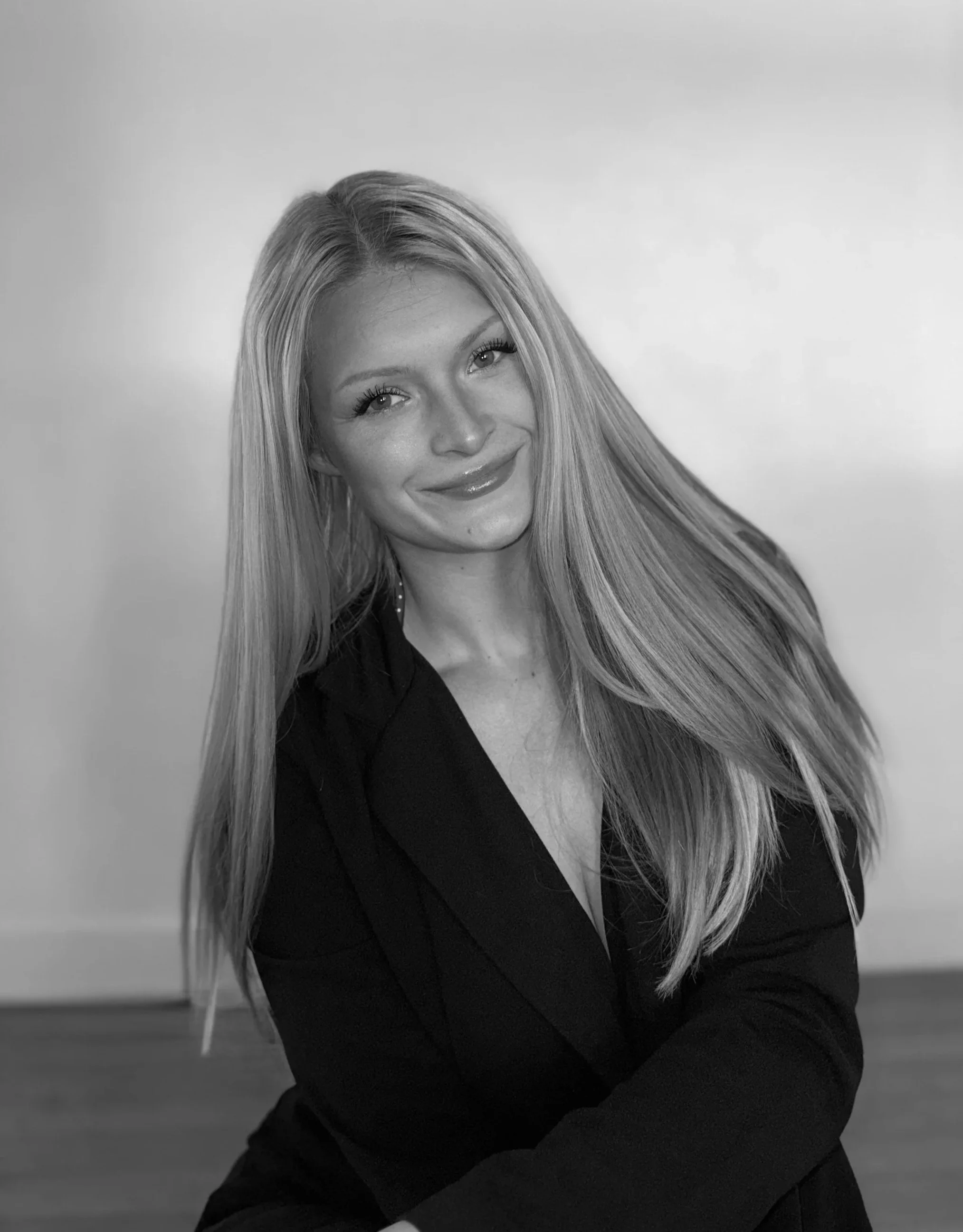 Black and white photo of a smiling woman with long, straight hair, wearing a dark blazer, posed indoors against a plain wall.