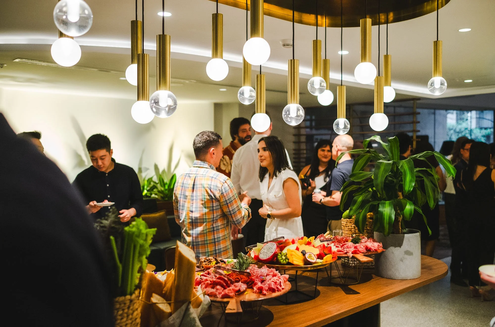 Guests mingling at a social gathering around a table with fresh fruit, vegetables, and meats, with modern hanging light fixtures overhead.